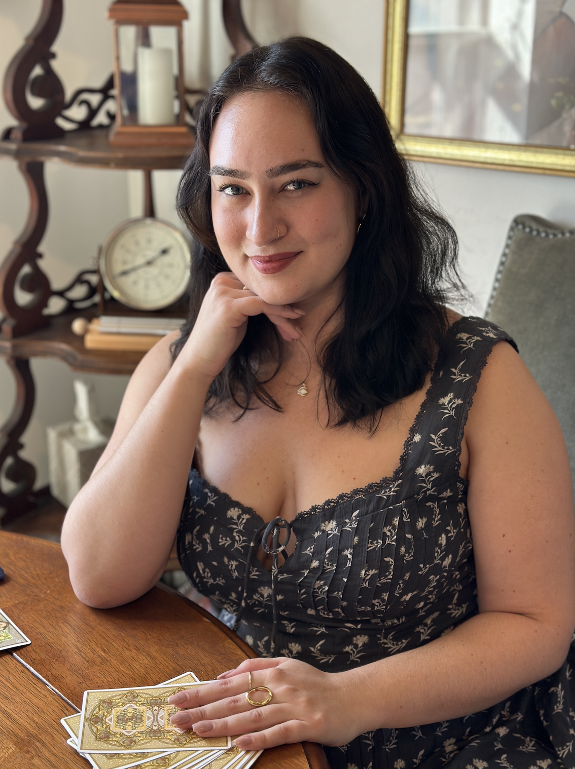 Black hair woman with grey navy dress sits at a wood table with one hand on tarot deck and the another hand on her chin