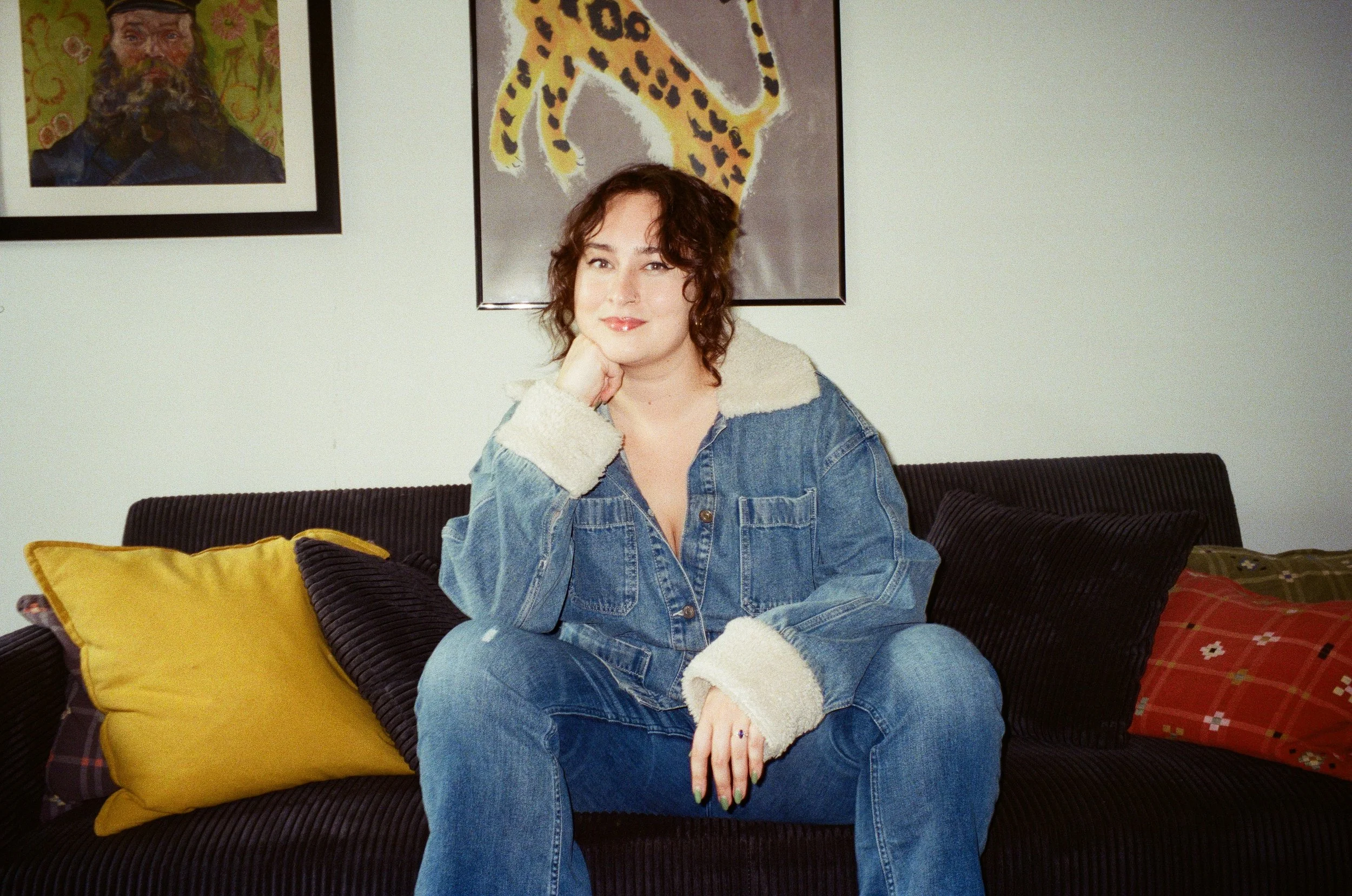 Brunette woman in jean jacket and jean pants sits on a black couch with one hand on her chin.