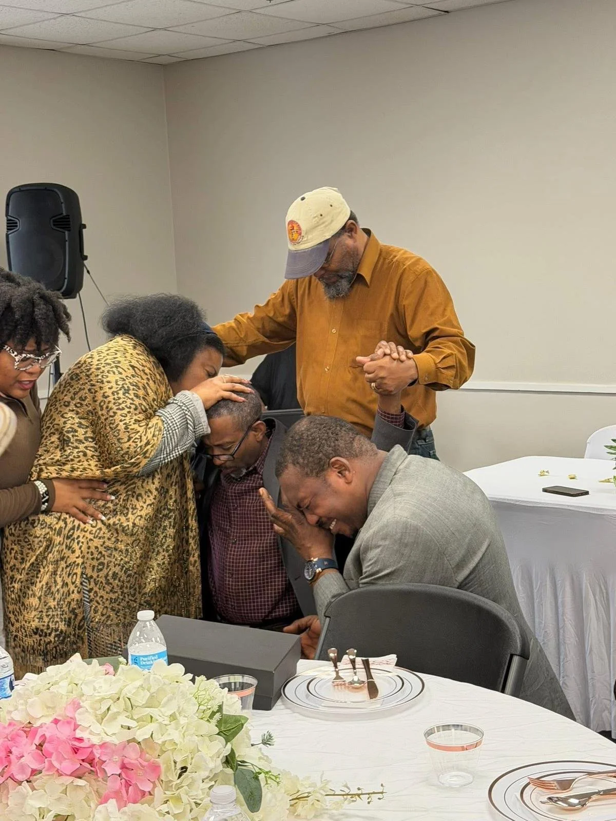 A group of people praying together at a celebration or gathering in a restaurant or banquet hall, with white tablecloths, flowers, and table settings visible.
