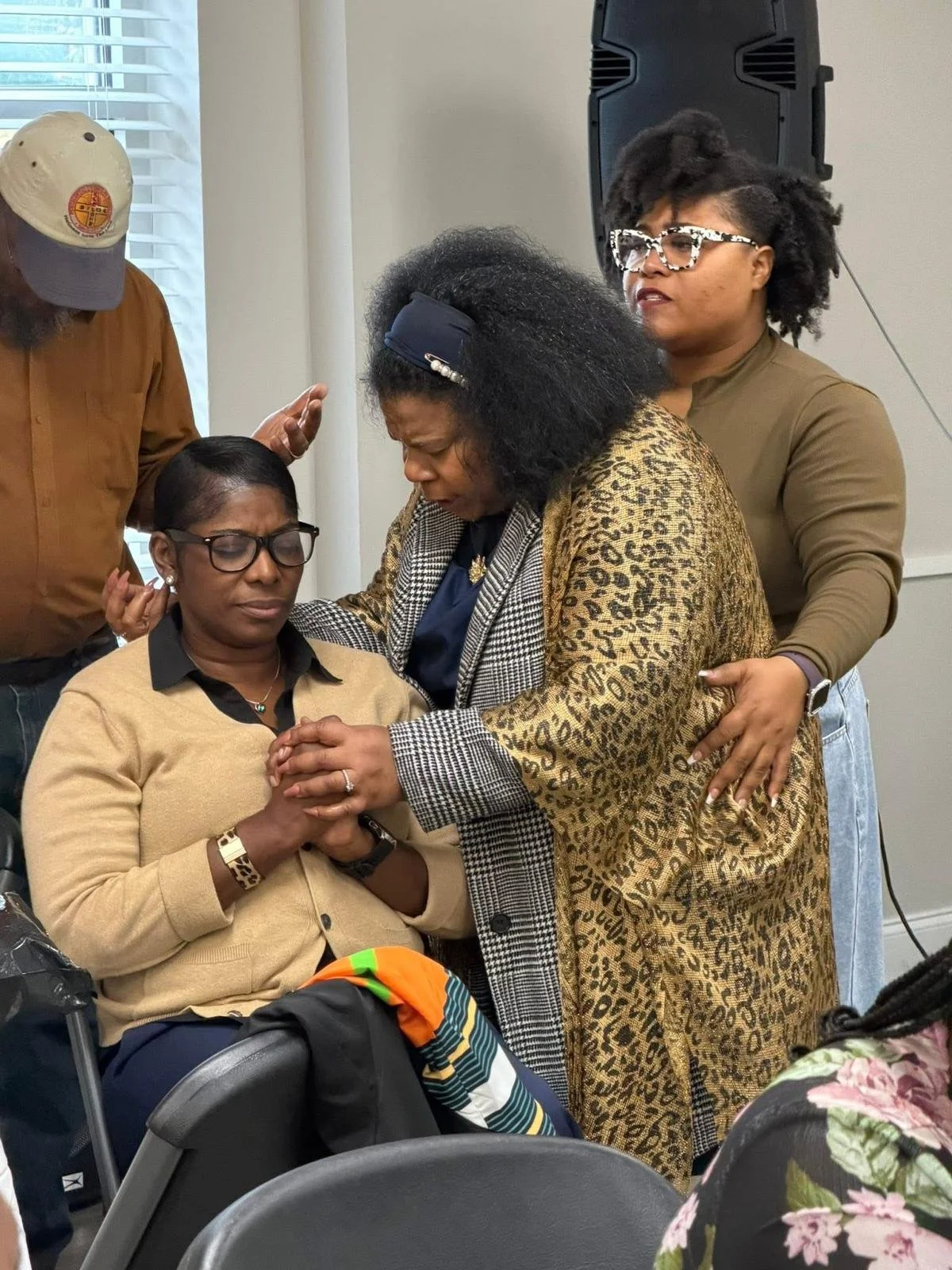 A group of women are praying together, with one woman seated with her hands clasped and eyes closed, while others stand around her, holding her hands or touching her shoulder in a supportive manner.