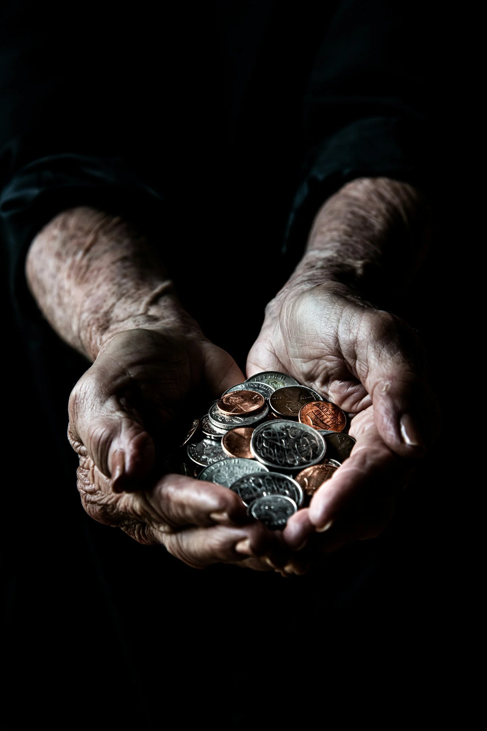 Close-up of an elderly person's hands holding a handful of mixed coins, including pennies, nickels, dimes, and quarters, against a dark background.