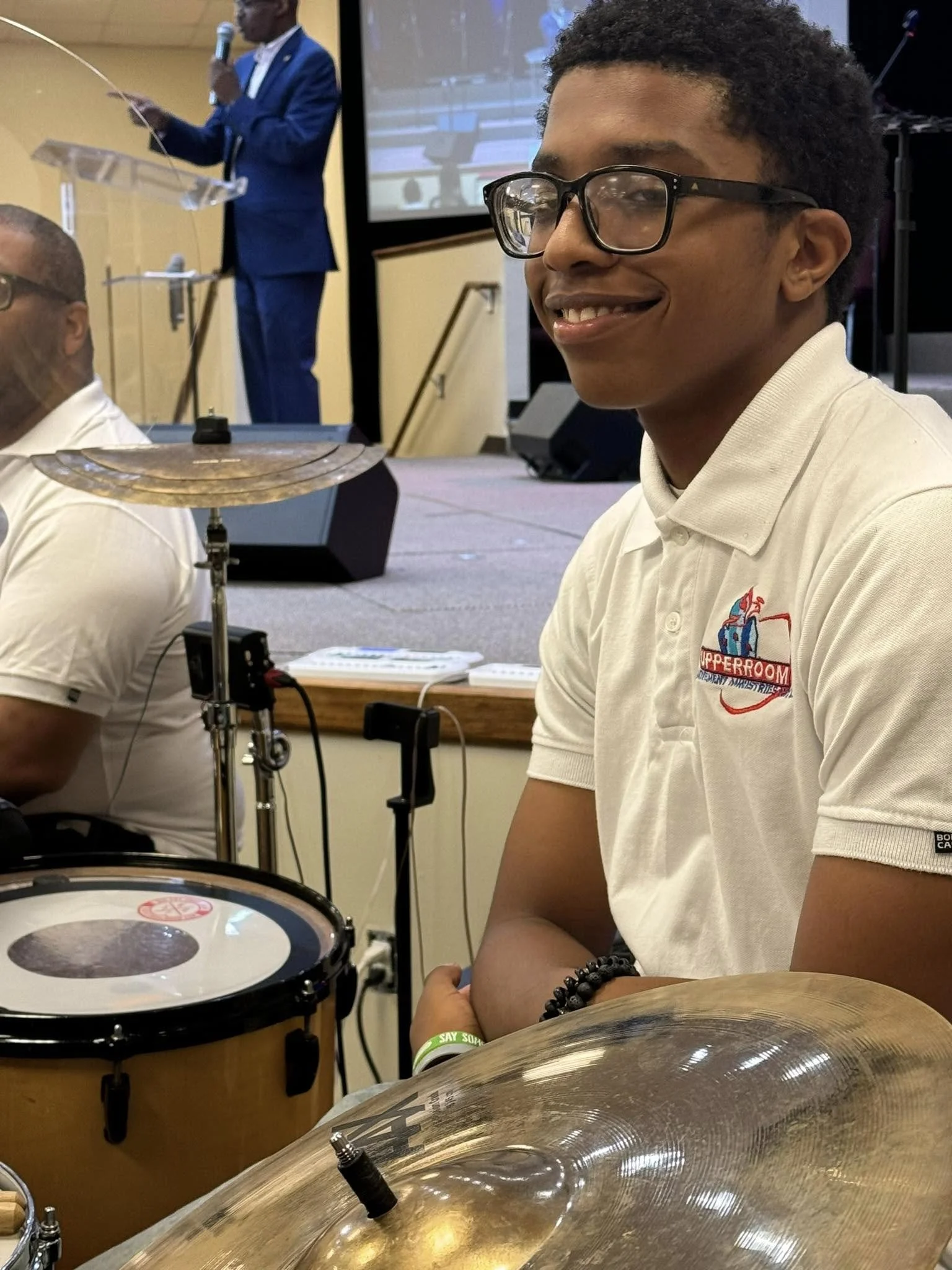 Young man with glasses smiling, sitting at a drum kit, wearing a white polo shirt with a logo, and a black beaded bracelet, at a music event.