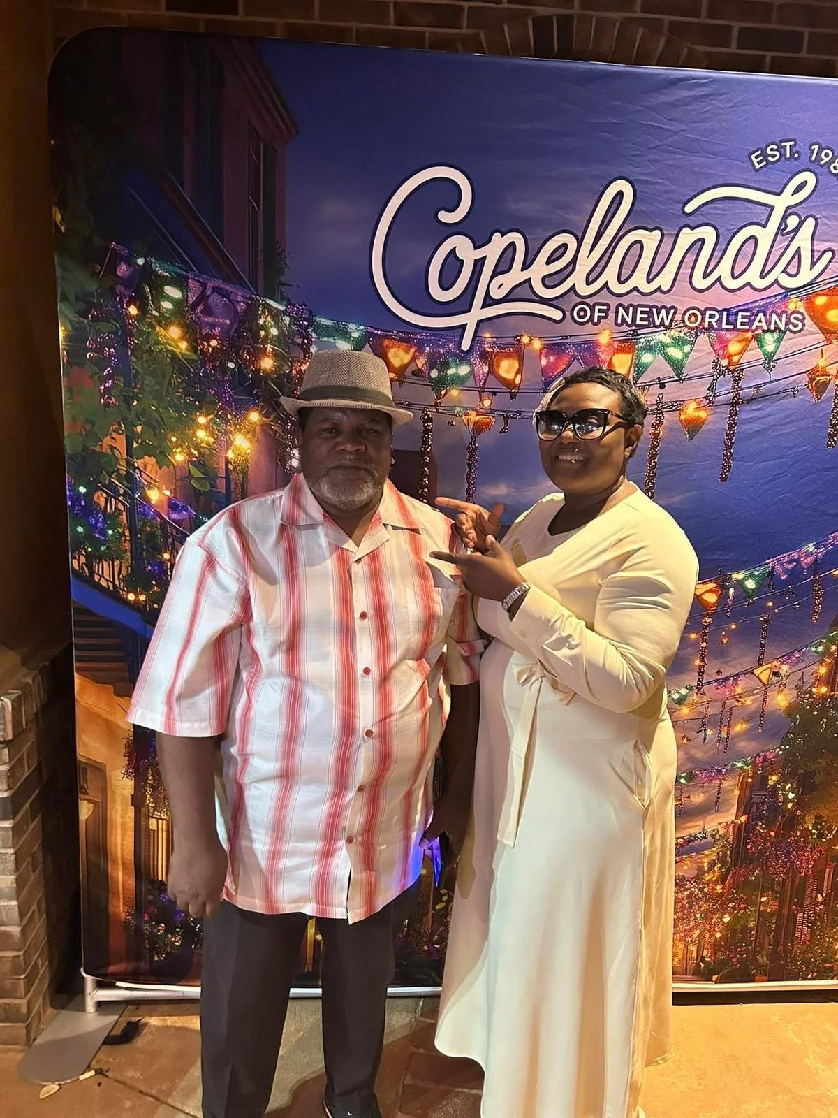 A man and woman standing together in front of a colorful New Orleans themed backdrop with string lights and the text 'Cafeland's of New Orleans'. The man is wearing a striped shirt and a fedora hat, and the woman is dressed in a light-colored dress with large sunglasses, pointing at the man.