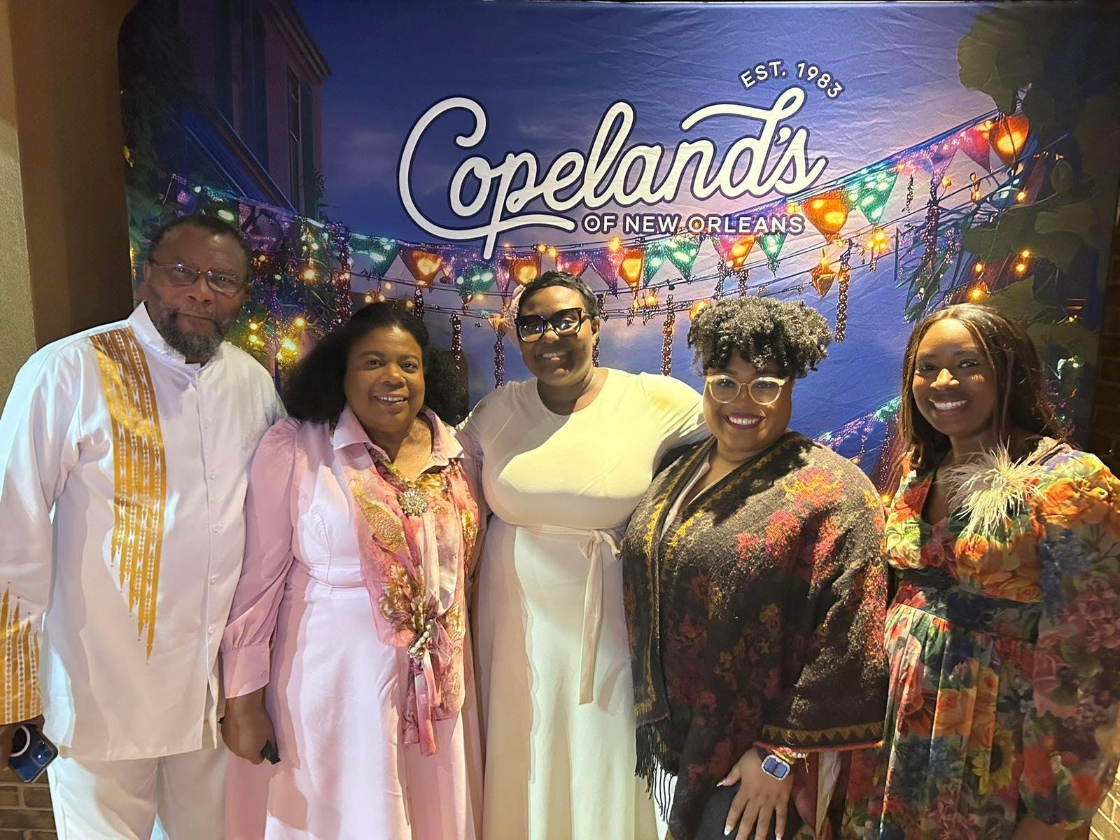 Six people standing together in front of a colorful Copeland's of New Orleans banner, smiling and celebrating.