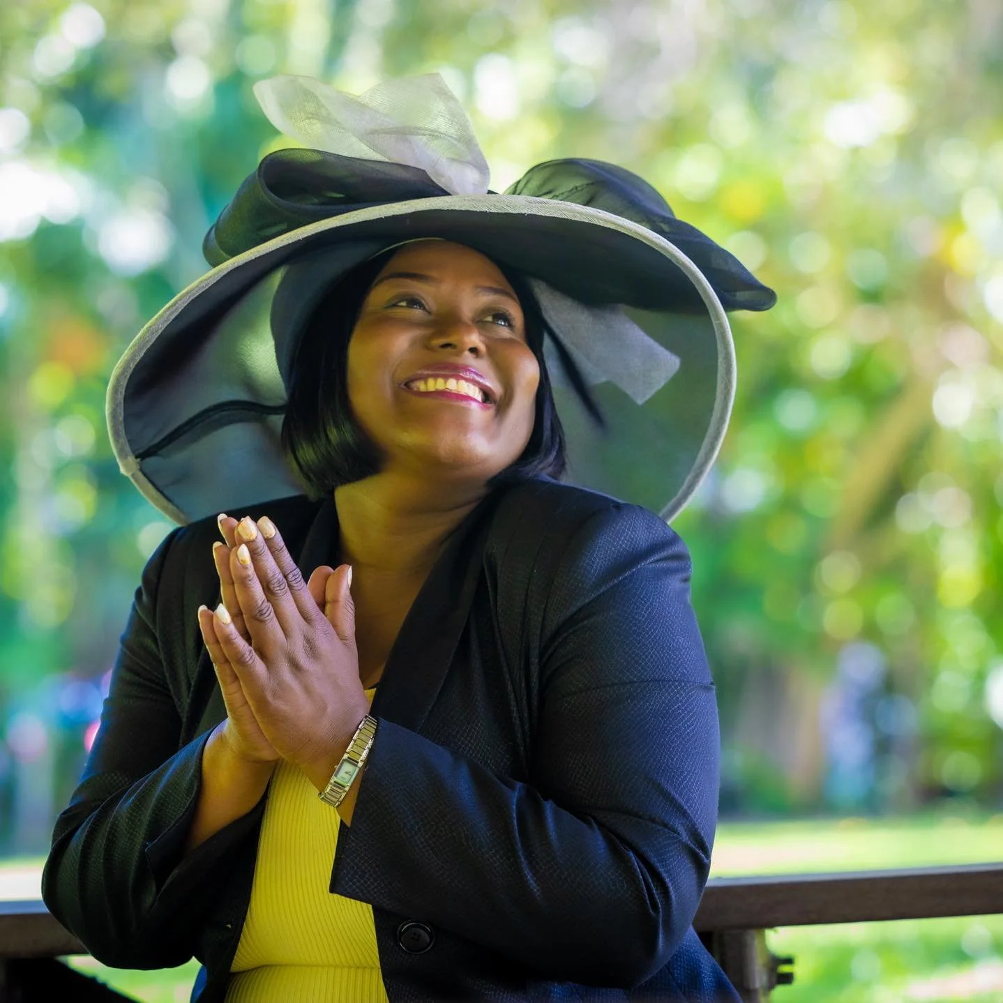 A woman wearing a large, black, wide-brimmed hat with a decorative bow and veil, smiling and holding her hands together near her chest, outdoors with a blurred green background.