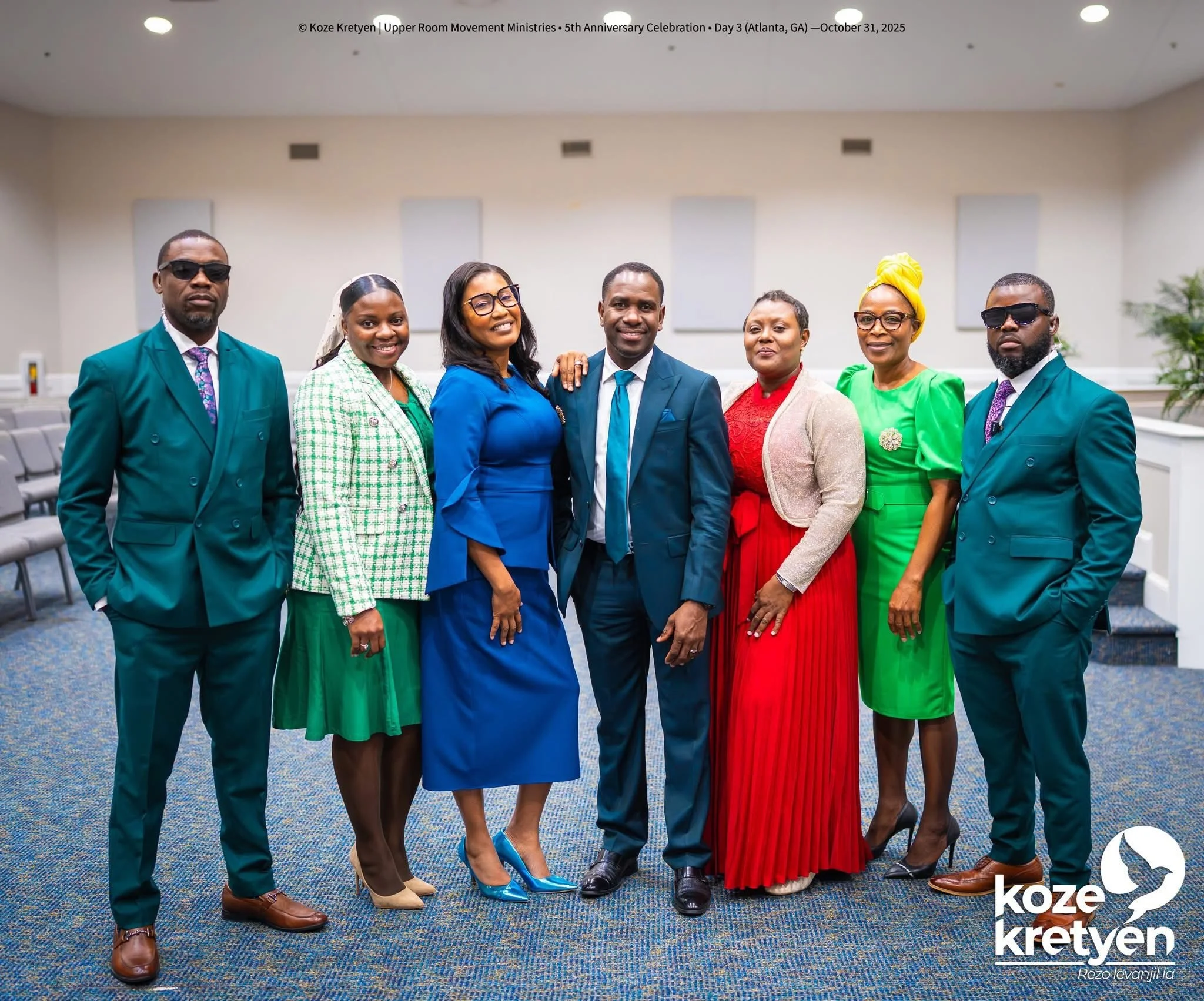 Group of seven diverse people dressed in vibrant and formal clothing, standing arm in arm in a conference room during an event. Four men and three women, some wearing glasses, with two men wearing sunglasses, smiling at the camera.