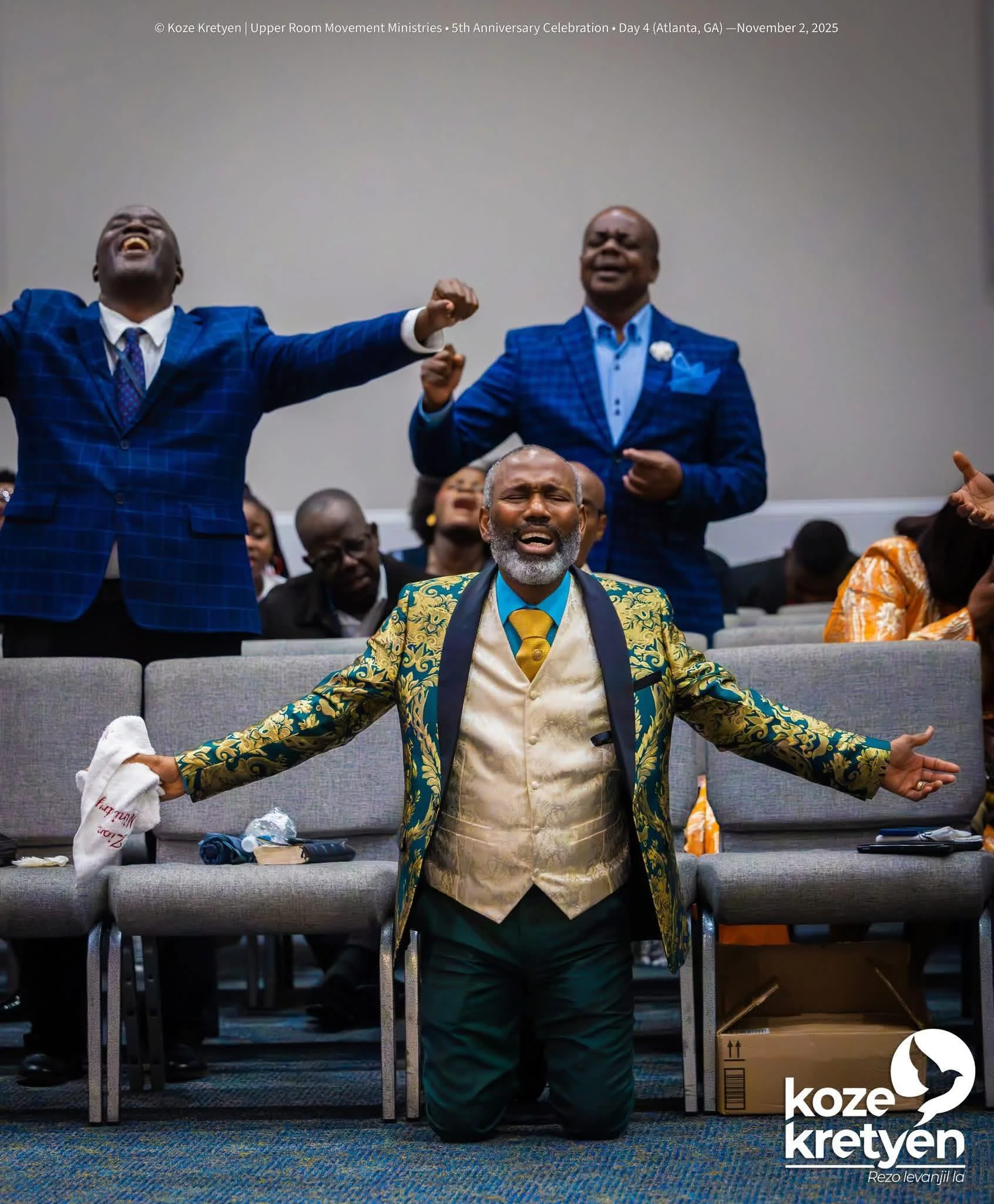A man in a patterned gold vest and ornate green jacket kneels with his arms outstretched in a church or conference room, with eyes closed and a joyful expression, while people behind him on stage are singing or praying, some with eyes closed. The setting appears vibrant and spiritual, with a mix of men and women dressed in formal and traditional clothing.