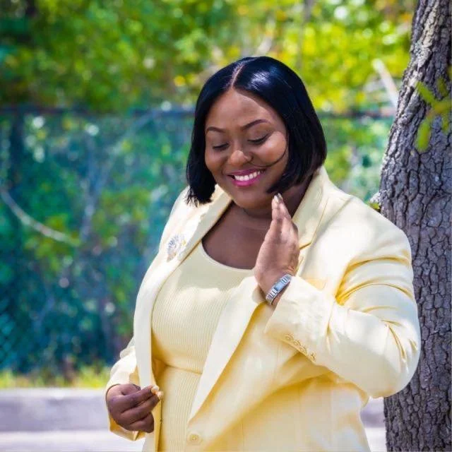 A woman with short black hair, wearing a yellow blazer and matching yellow top, standing outdoors next to a tree with green leaves in the background, smiling and looking down.