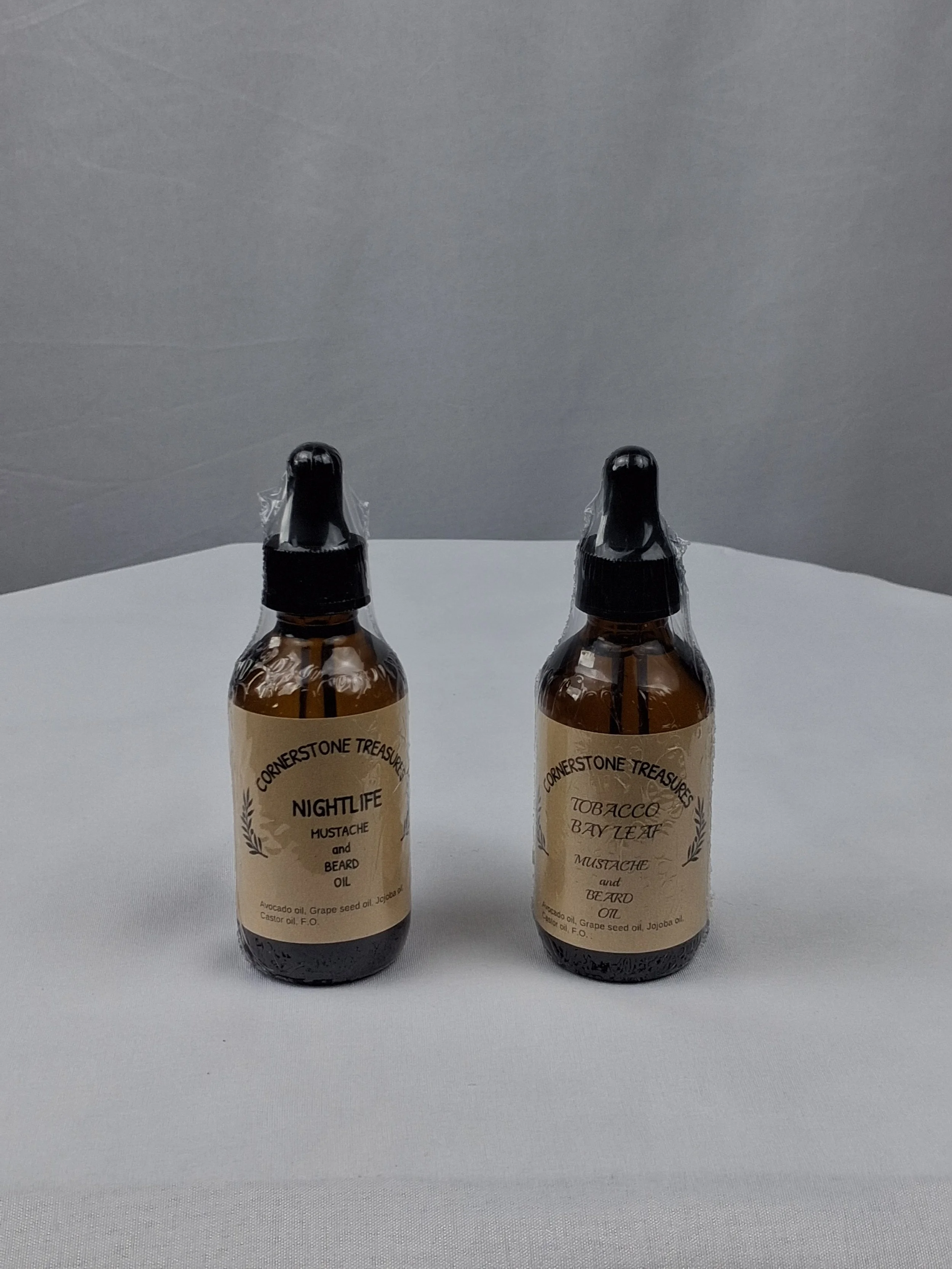 Two brown bottles of Cornerstone Treasures Nightlife Mustache and Beard Oil on a white surface with a gray background.