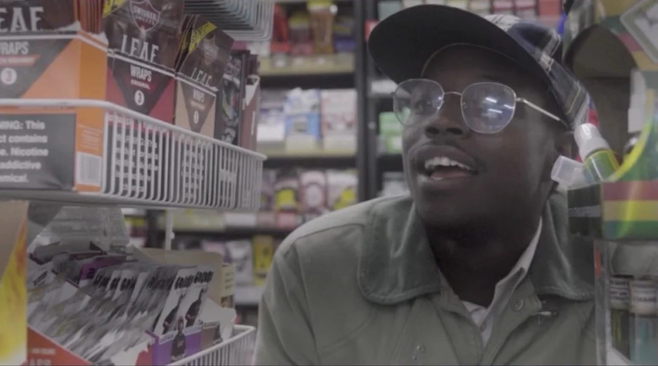 A man wearing glasses, a baseball cap, and a jacket, looking at products on store shelves in a grocery store aisle.
