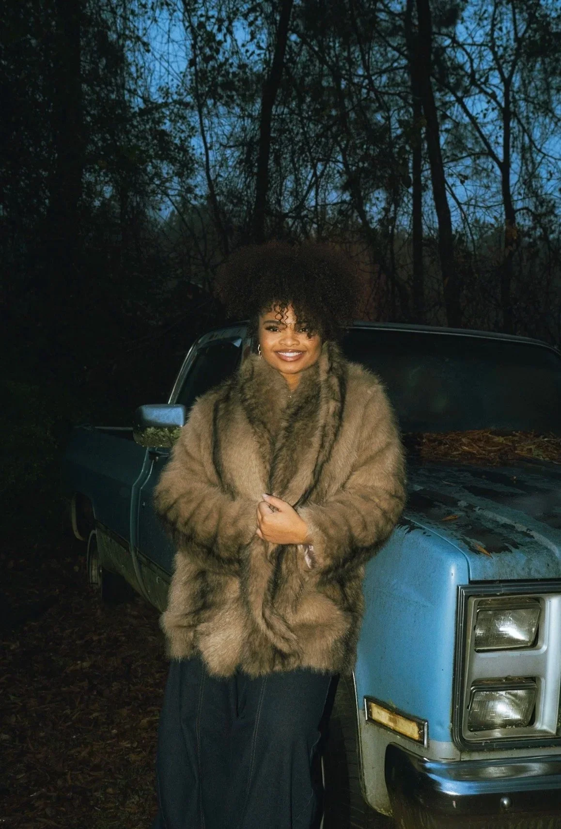 A woman with curly hair smiling, wearing a fur coat, standing in front of a blue pickup truck in a wooded area at dusk.
