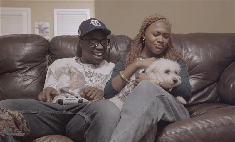 A man and woman sitting on a brown leather couch with a small white dog, the woman is petting the dog and the man is smiling.