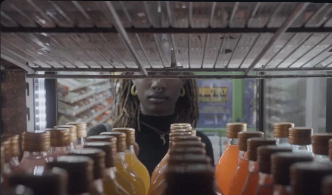 A person with dreadlocks and gold jewelry looking at the camera through a store shelf filled with colorful bottles of beverages.