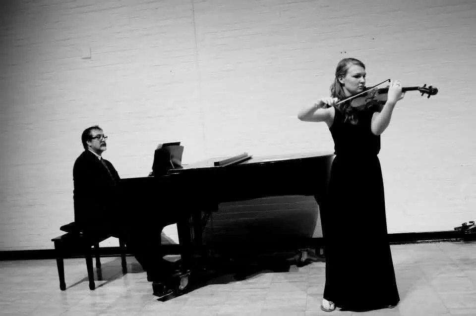 A woman in a black dress playing the violin while a man in a suit plays the piano in the background.