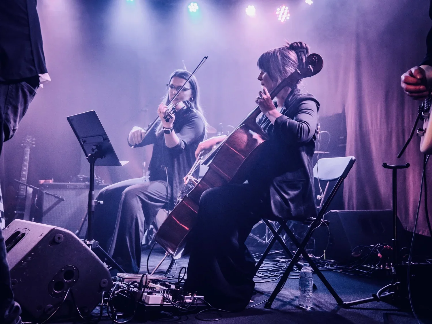 Two women playing musical instruments on stage, one with a violin and the other with a guitar, during a live music performance with purple stage lighting.