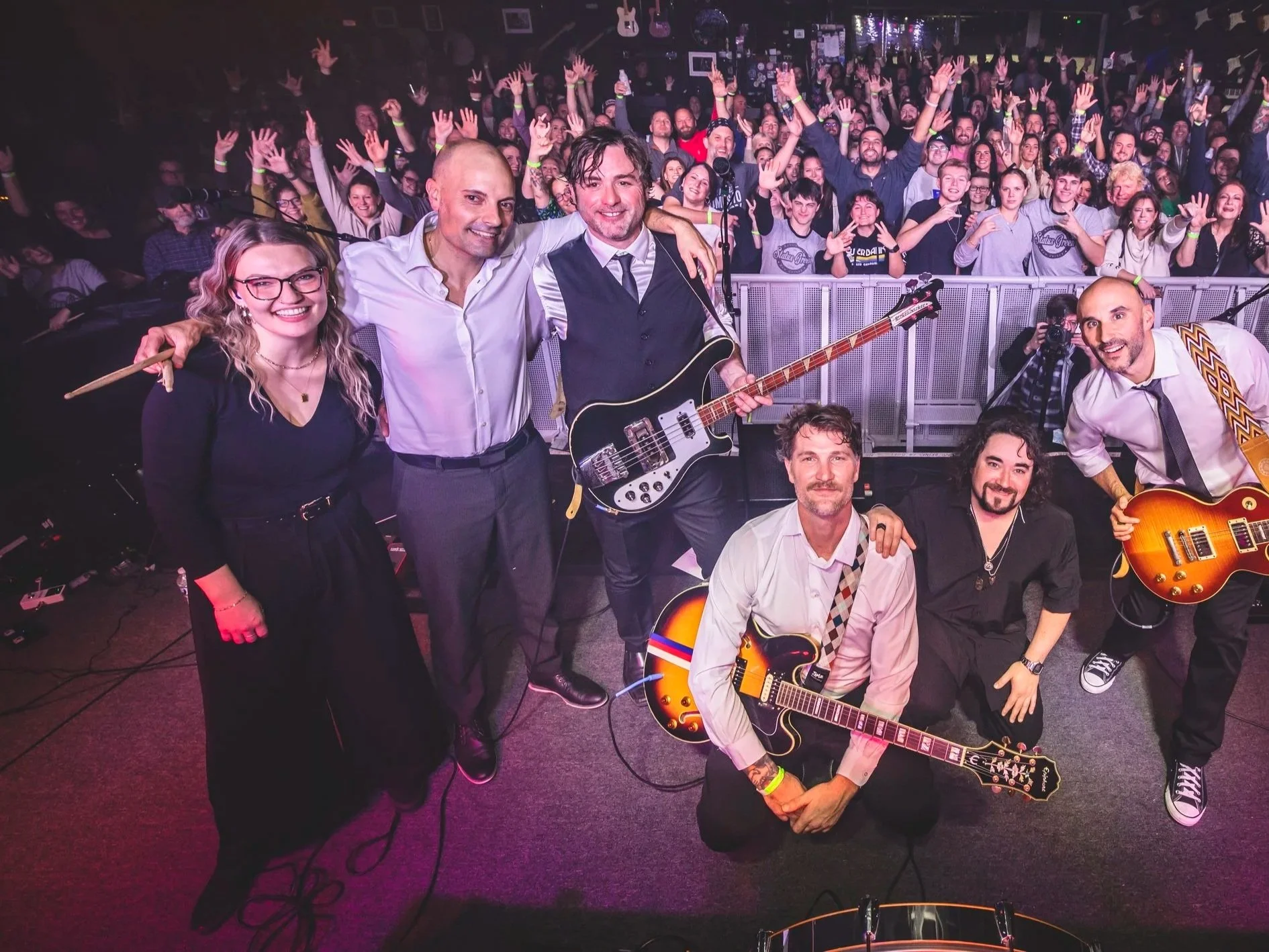 A band of musicians taking a selfie on stage with a large crowd of concertgoers raising their hands in the background inside a music venue with guitars hanging on the wall and reflective disco balls hanging from the ceiling.
