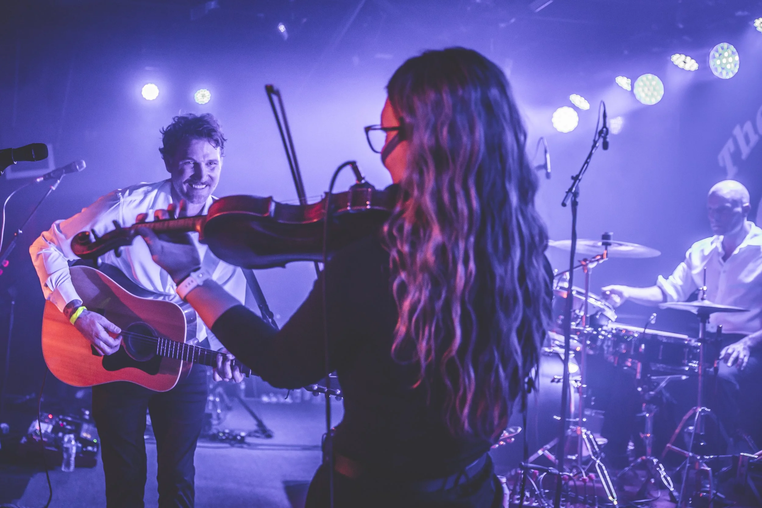 A woman with long, curly hair and glasses stands in front of a male guitarist playing an acoustic guitar during a live performance. The background features stage lights and music equipment.