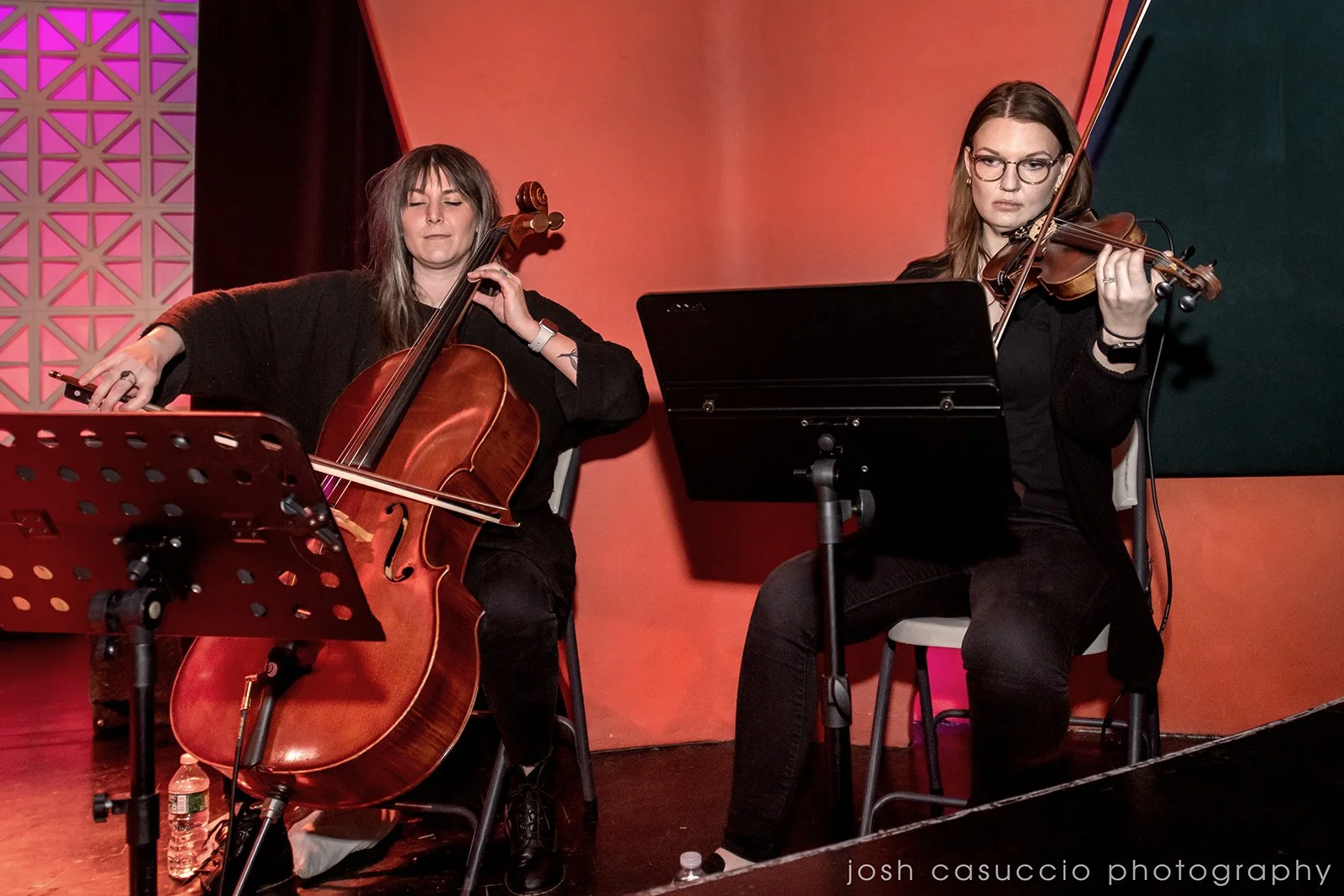 Two women playing classical instruments, a cello and a violin, during a live music performance in a cozy indoor setting.
