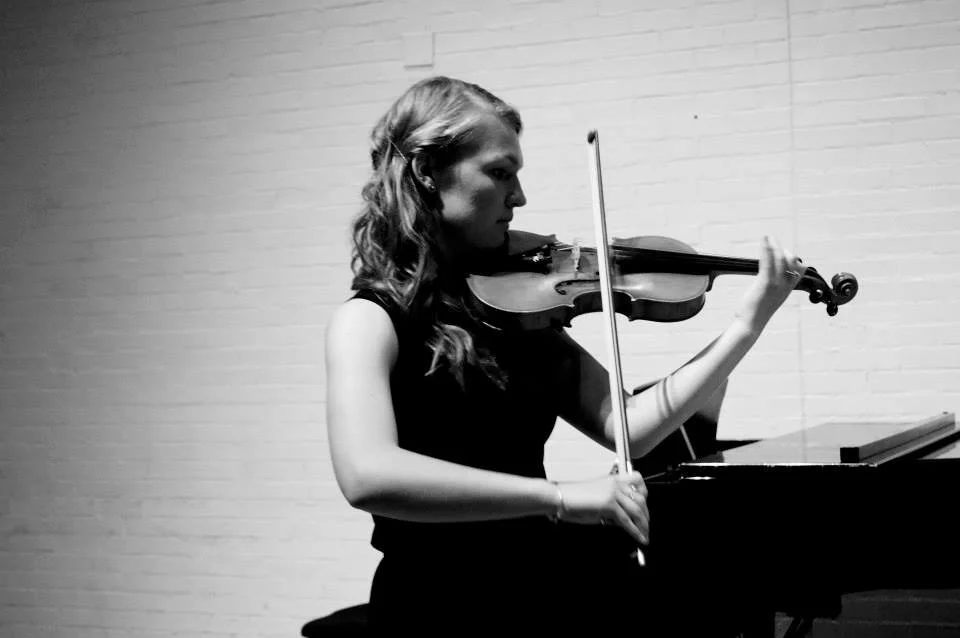 A young woman plays the violin at a piano, standing against a white brick wall.