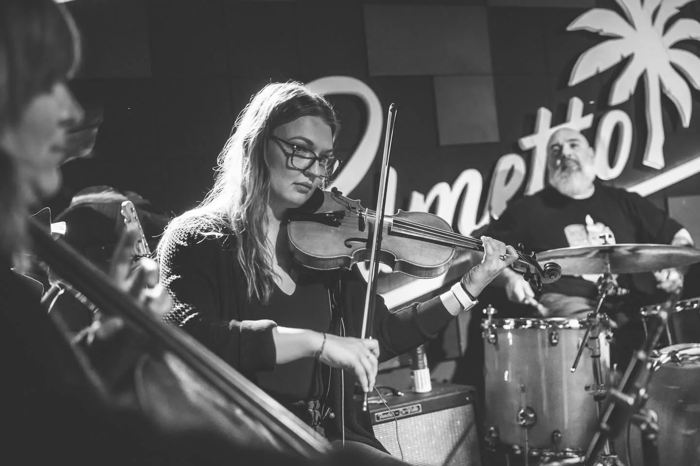 Black and white photo of a woman playing the violin with a man playing drums in the background at a live music venue.