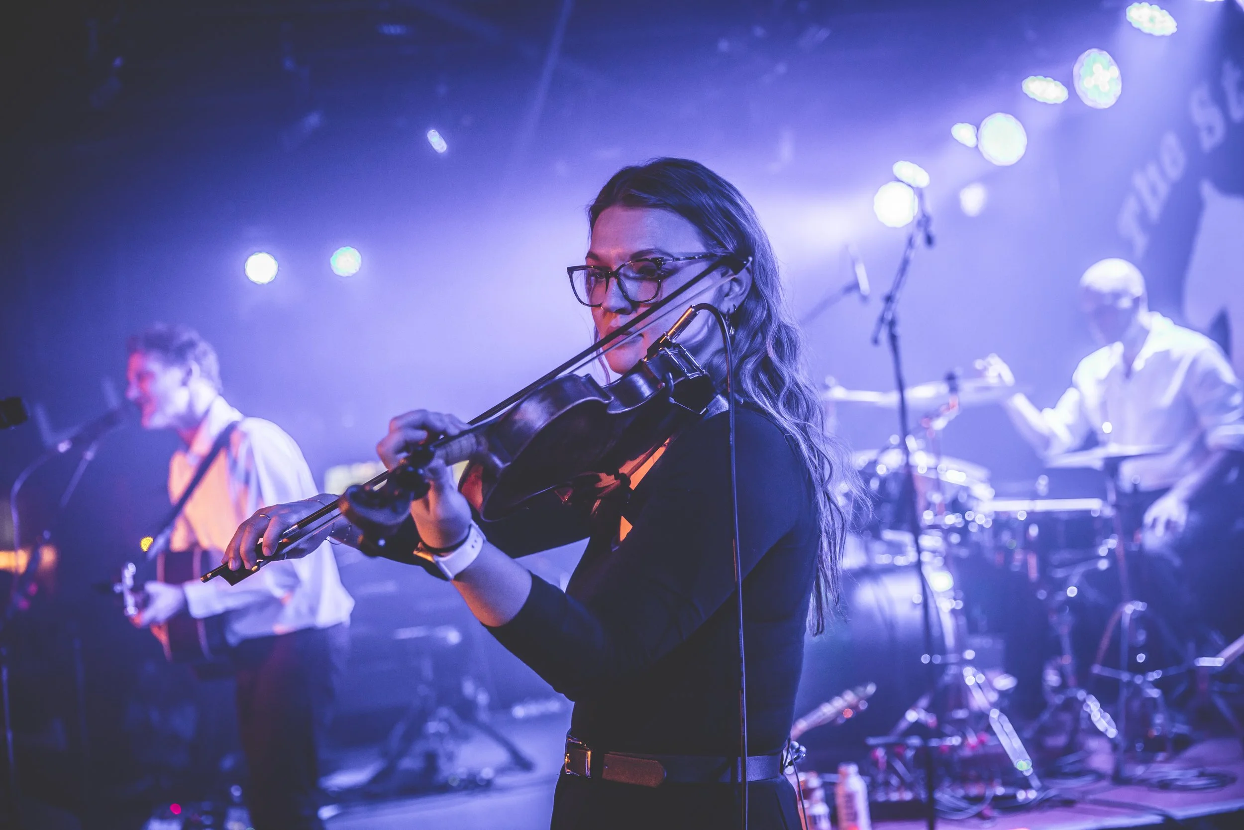 Haley S playing the violin on a stage with blue and purple lighting, with other musicians visible in the background.