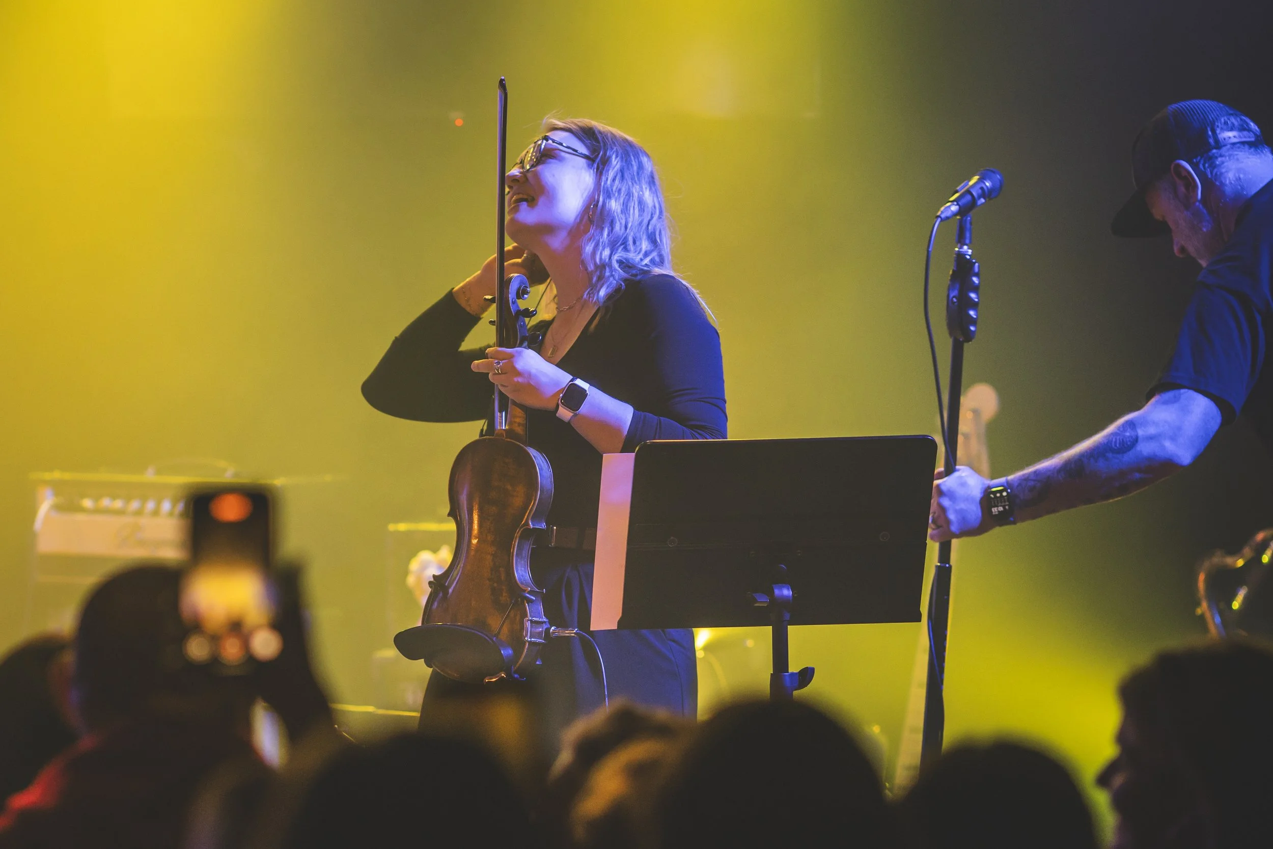 Haley Schilla playing a violin on stage, smiling and wearing glasses, with a man adjusting sound equipment nearby, in a concert setting with yellow and green lighting. All Rights Reserved.