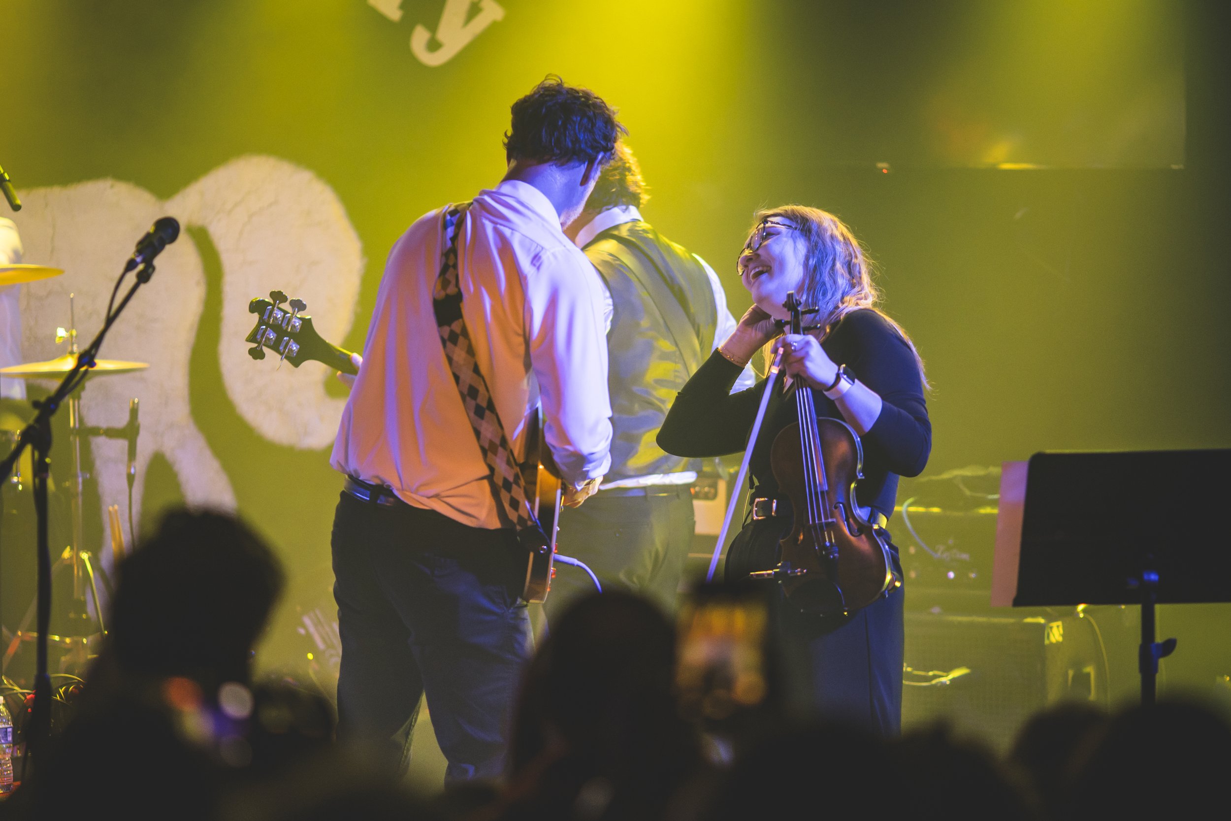 A group of musicians on stage, with a woman playing the violin and smiling at two men holding guitars, under yellow stage lighting.