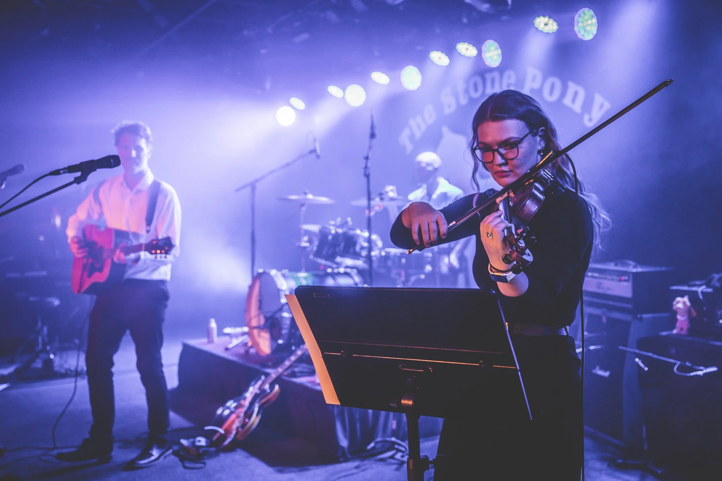 A woman playing the violin on stage, with band members in the background, illuminated by blue and purple stage lighting at a live music venue.