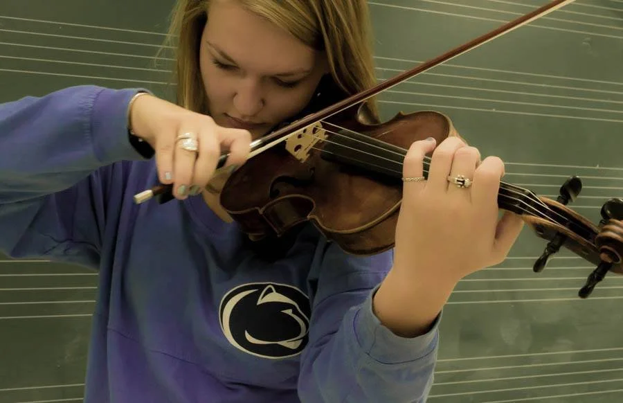 Young woman playing the violin, standing in front of a chalkboard with musical staff lines.