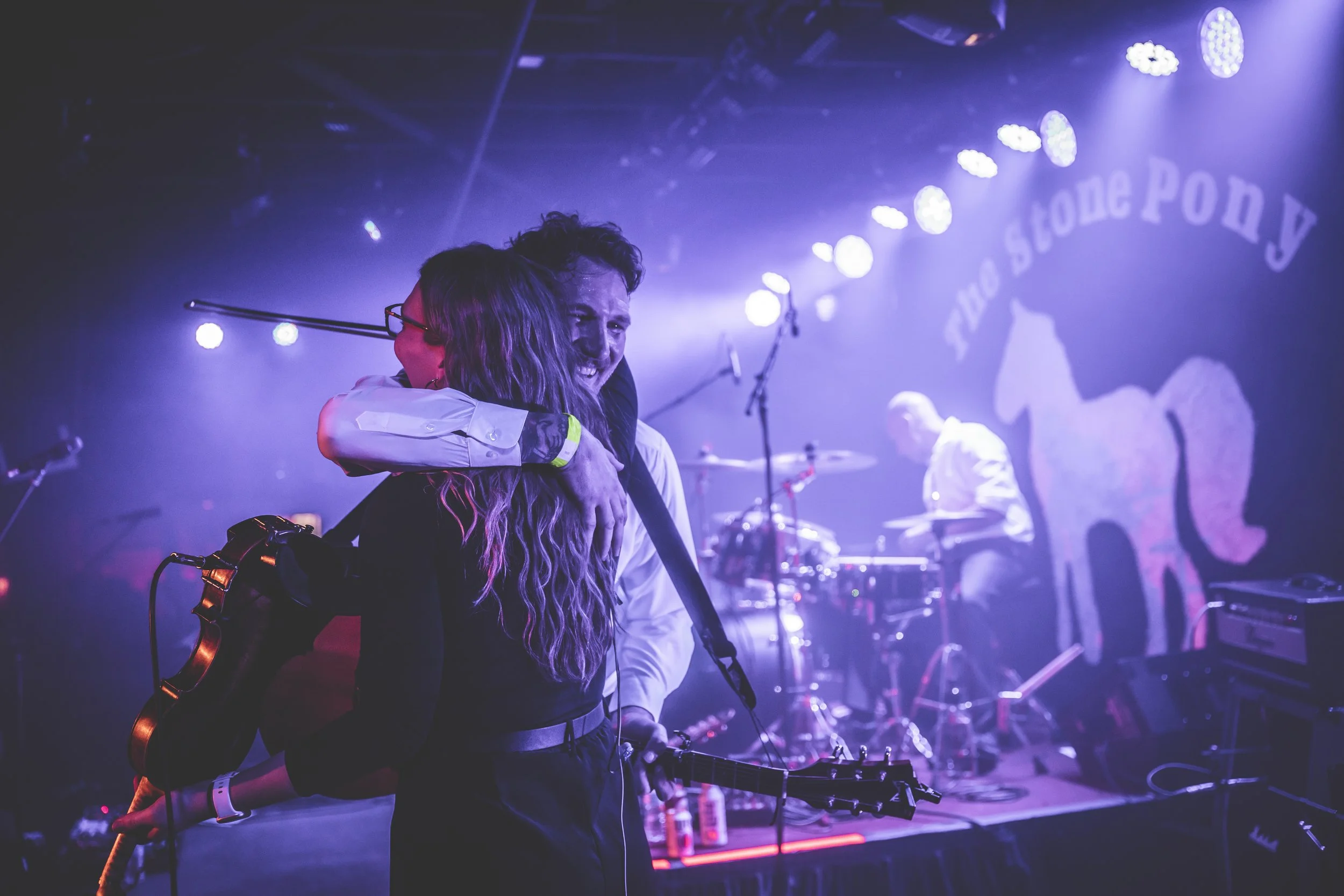 Two people hugging on stage during a live music performance, with band members and stage lights in the background.