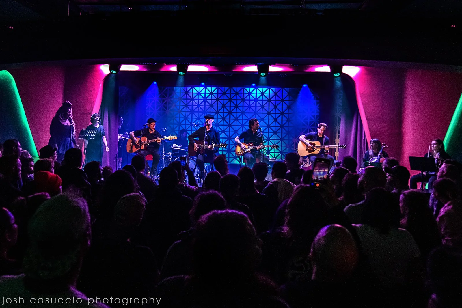 A live music performance featuring a band on stage with several musicians playing guitars and singing, illuminated by colorful stage lights, with an audience watching.