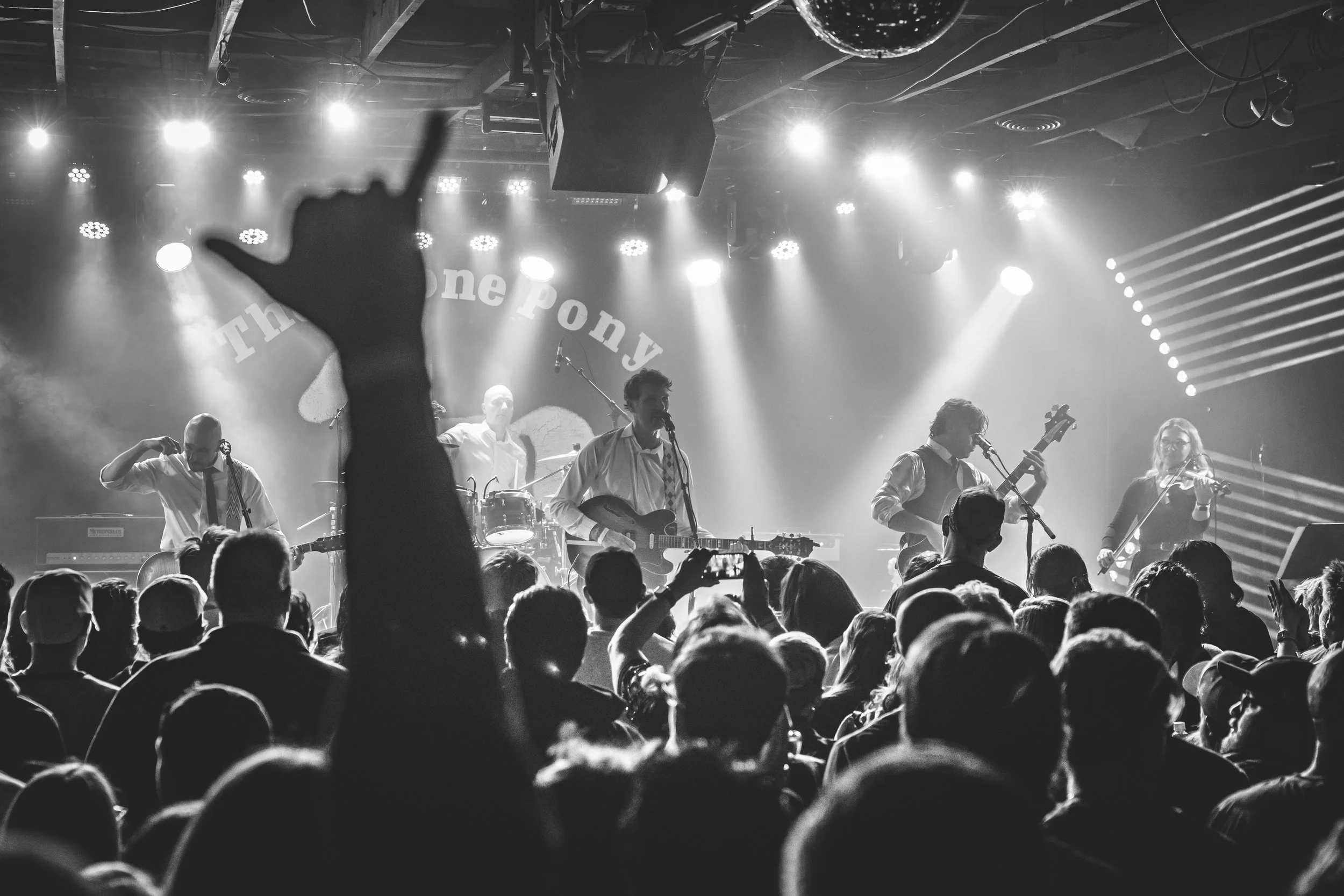 A live music band performing on stage at a concert with bright lights. The audience is visible in the foreground, with many people raising their hands and taking photos.