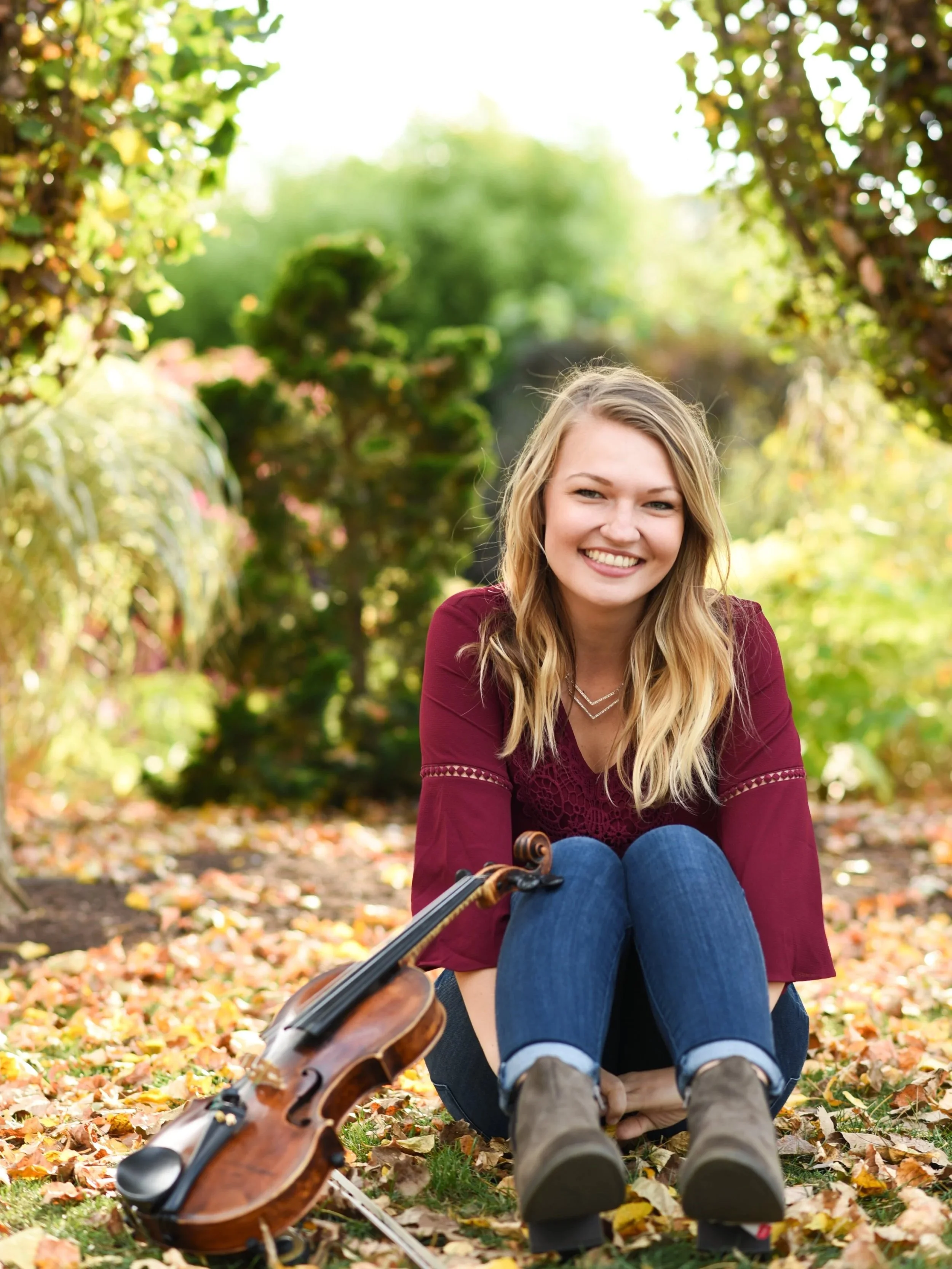 A young woman with long blonde hair sitting on the ground in a park covered with fallen autumn leaves. She is smiling and looking at the camera, holding a violin and bow, with a guitar lying in front of her.