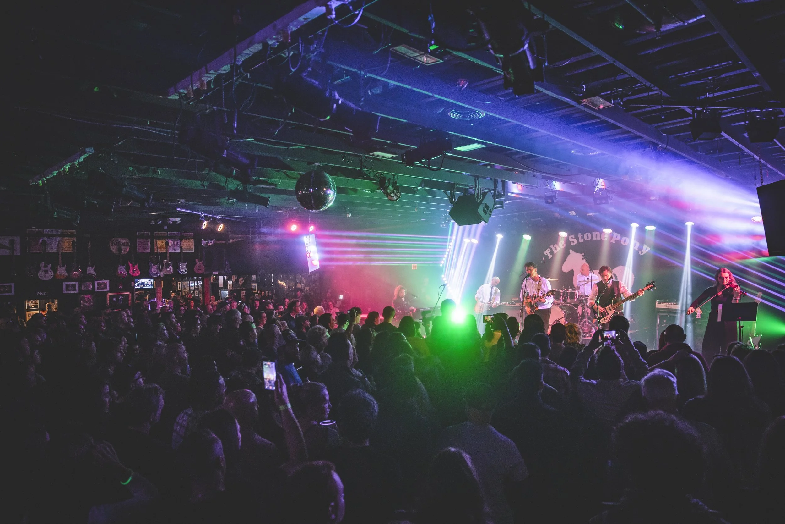Concert with band performing on stage, colorful lights, and an audience watching in a nightclub with guitars on the wall.