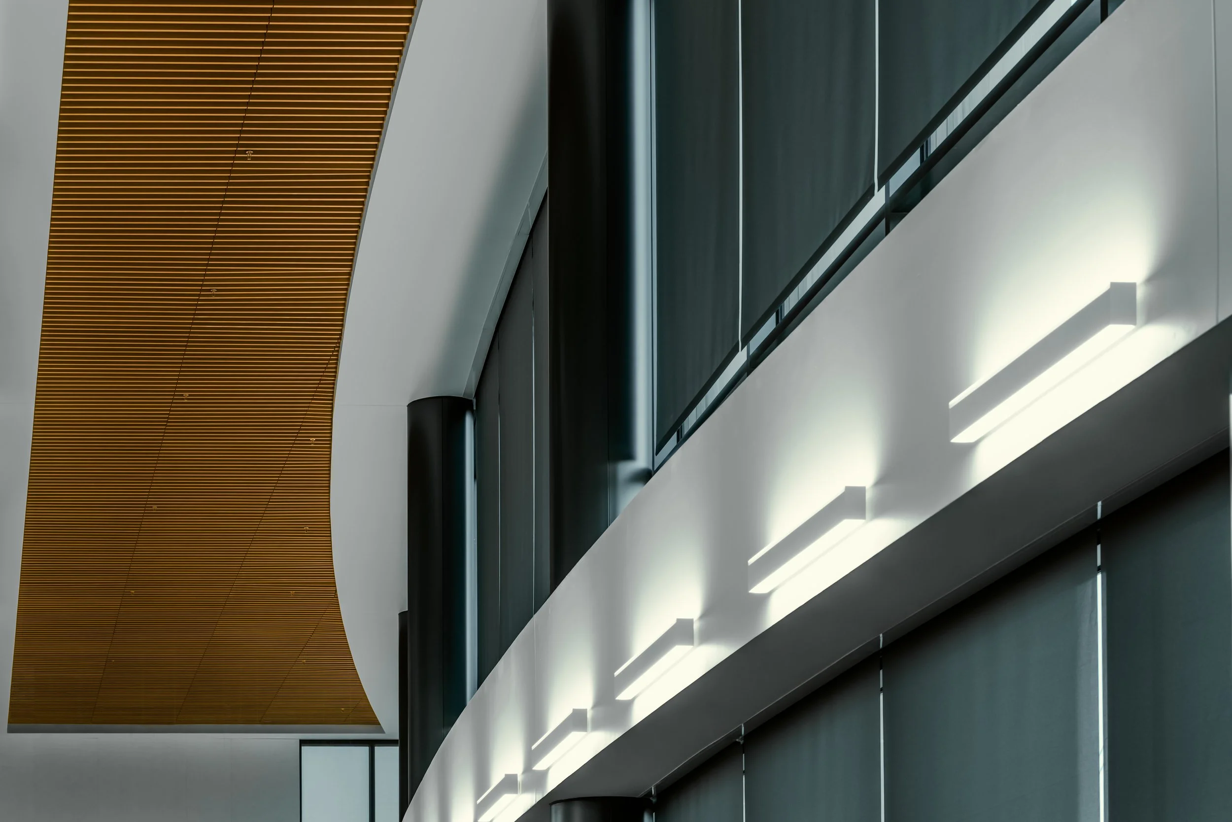 Interior of a modern building with angled ceiling, featuring a large yellow slatted wooden panel on the ceiling, gray walls, and sleek white lighting fixtures.
