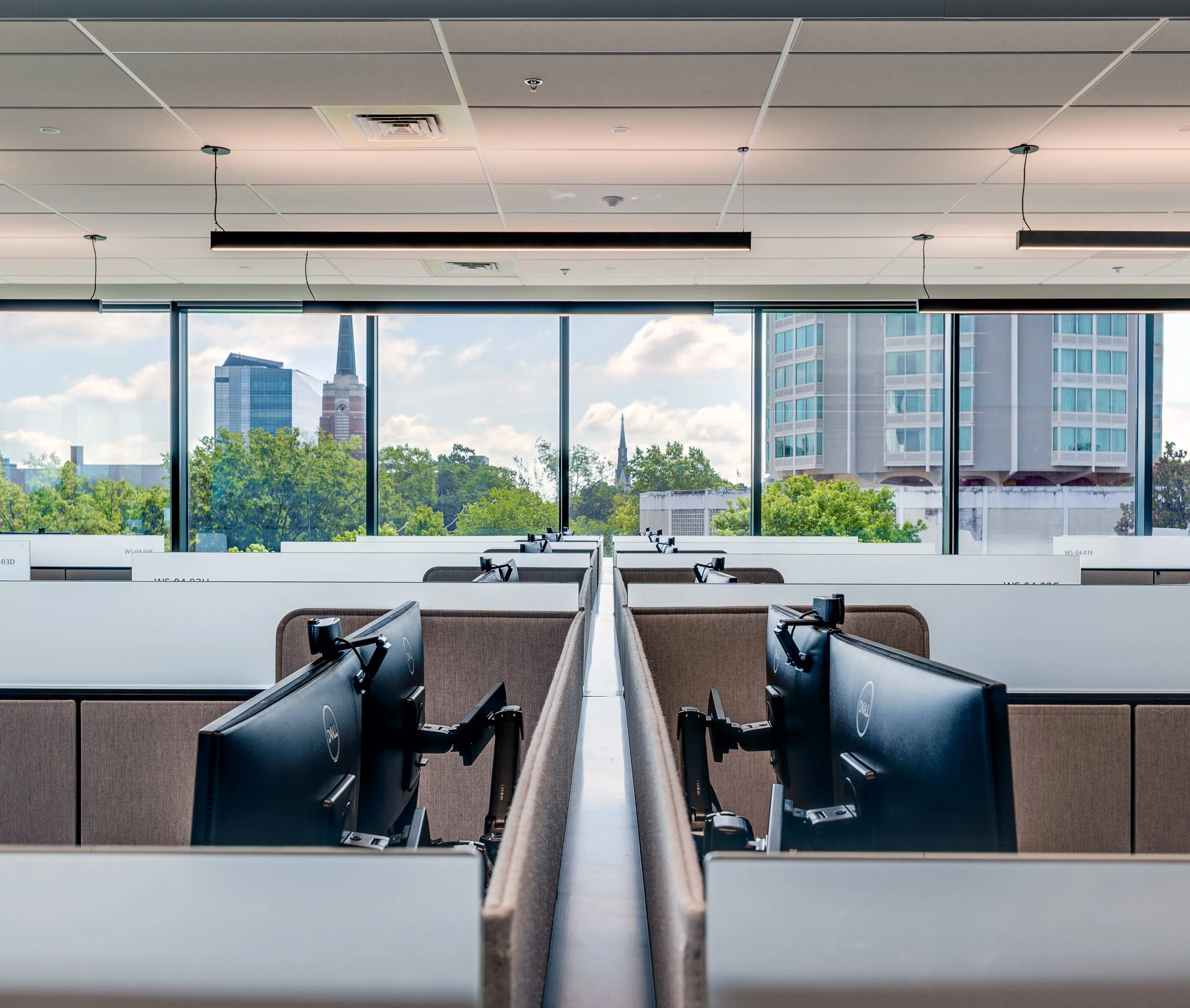 Office space with rows of cubicles equipped with computer monitors, large windows showing city buildings and blue sky outside, modern ceiling lights, and city skyline in the background.