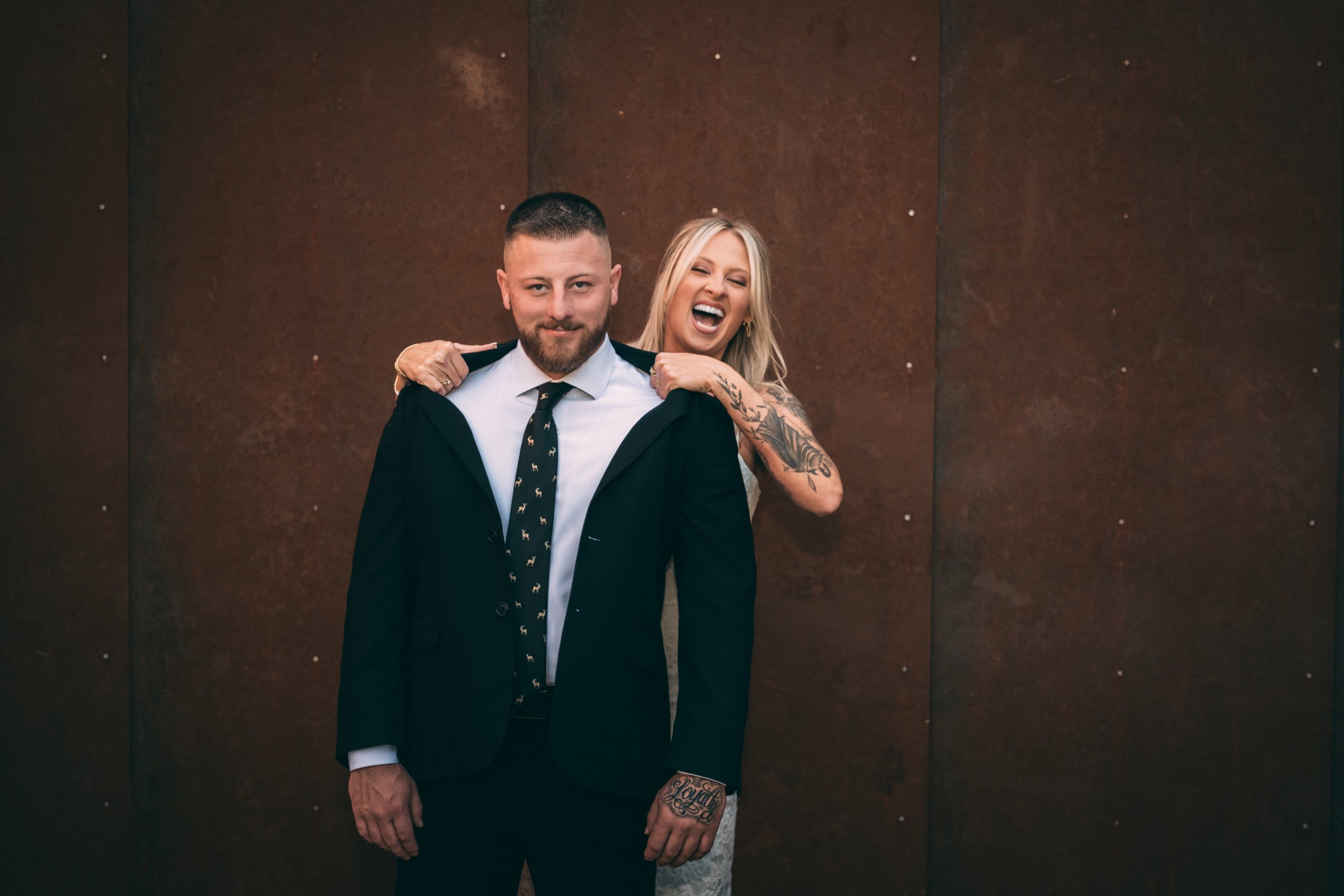 A woman with long blonde hair laughing and a man with short hair and beard, wearing a suit and tie, standing in front of a rust-colored wall.