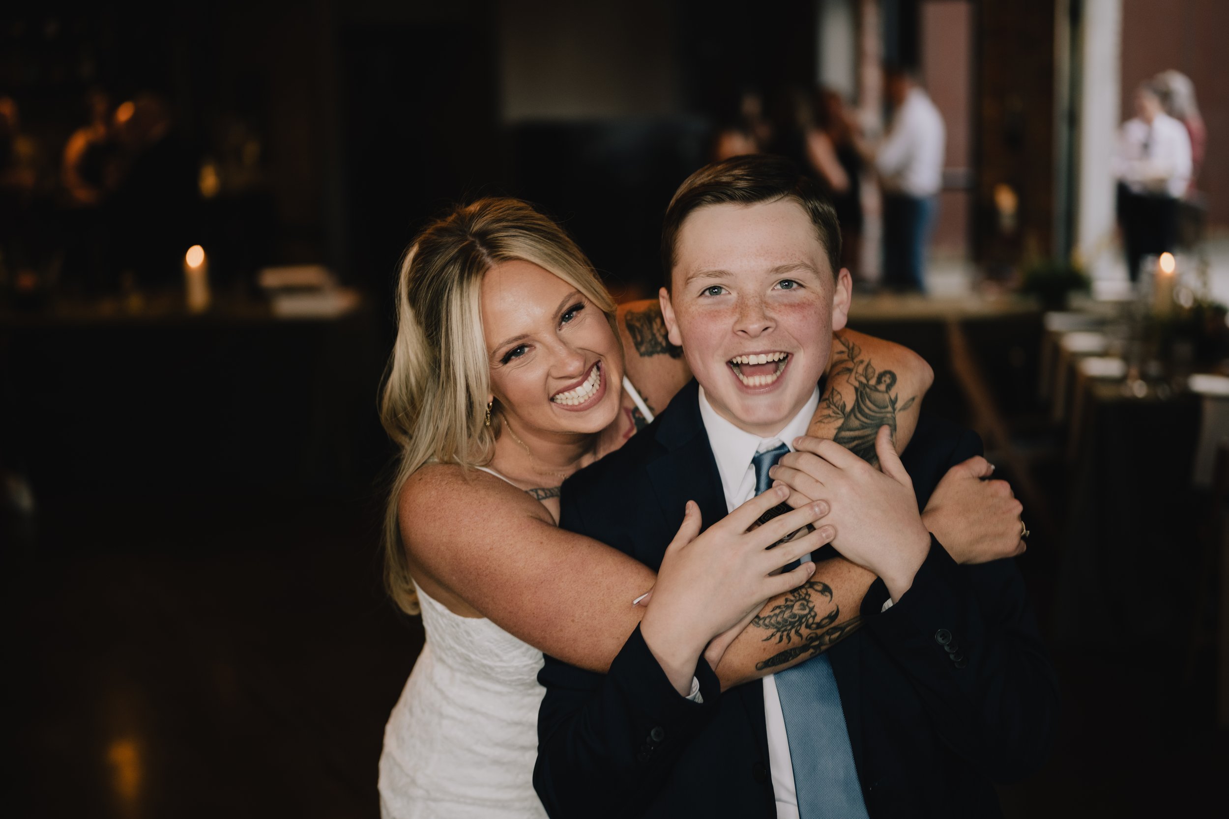 A woman with blonde hair and tattoos embraces a young boy in a suit at a social gathering or celebration, with people and dim lighting in the background.