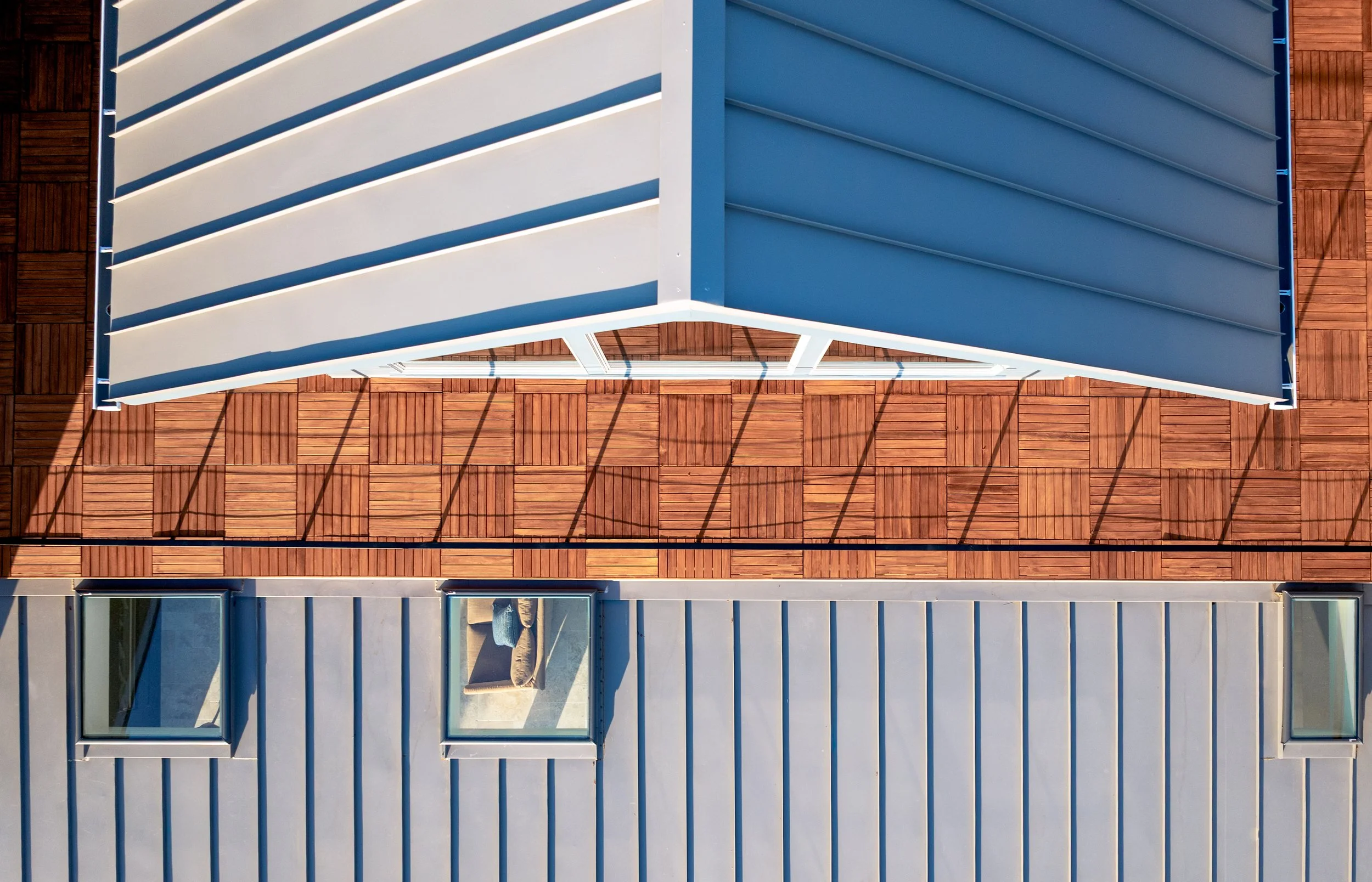 An aerial view of a modern building with a rooftop deck. The roof features white and blue horizontal slats, and a section with wooden tiles. There are large windows visible on the side of the building.