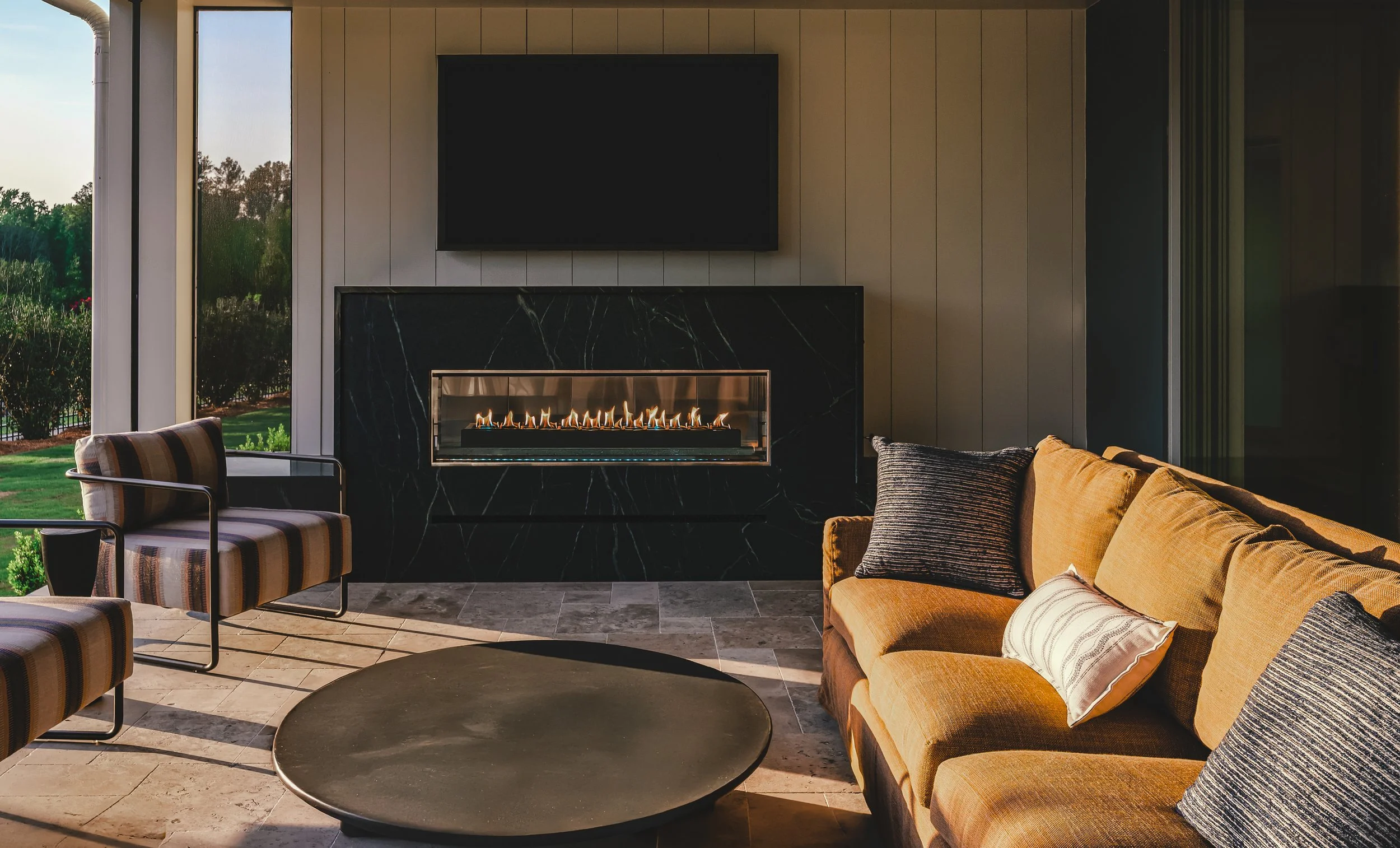 A cozy living room with a gas fireplace, a wall-mounted TV above it, a beige sofa with pillows, a round coffee table, and large windows showing a lush green outdoor landscape.