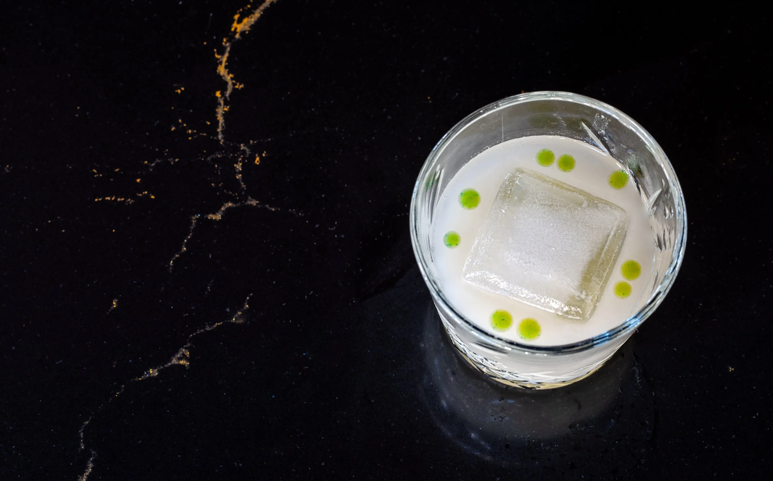 A glass of cocktail with a large ice cube and small green dots, placed on a dark black stone countertop with gold veining.