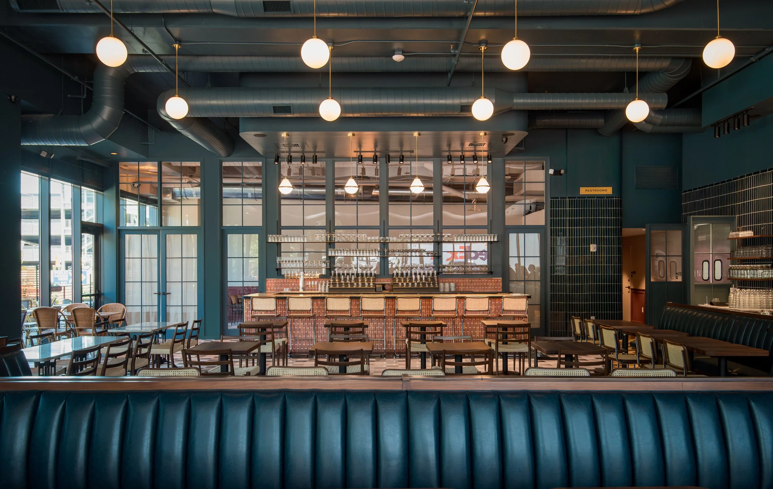 Interior of a modern restaurant or cafe with a brick bar counter, multiple wooden chairs and tables, large glass windows, hanging pendant lights, and exposed ductwork on the ceiling.