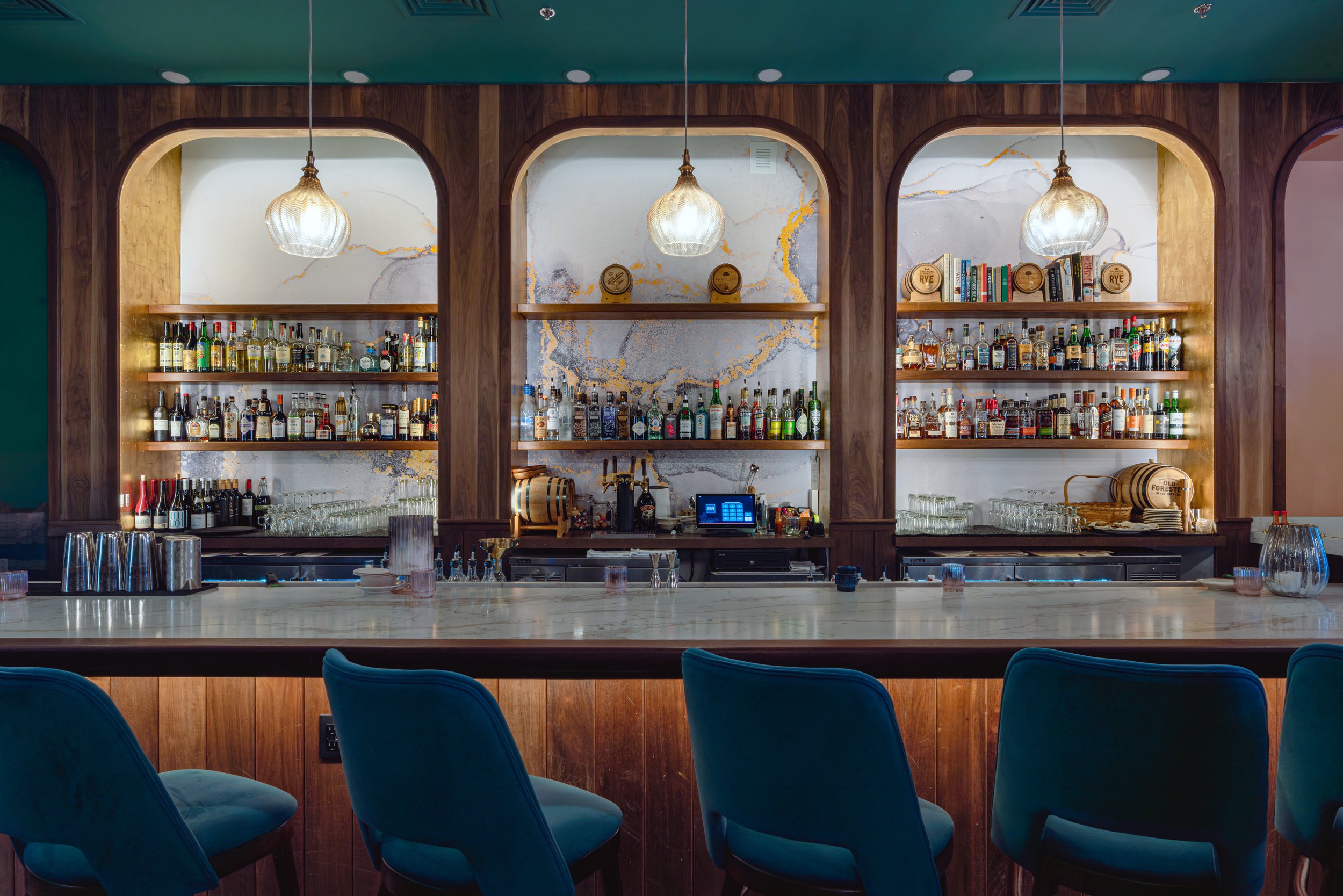 A bar with a marble countertop and a wooden back wall featuring shelves filled with various bottles of liquor. Above the bar, there are three decorative pendant lights hanging, and behind the bar, there is a cash register and glasses. Blue chairs are