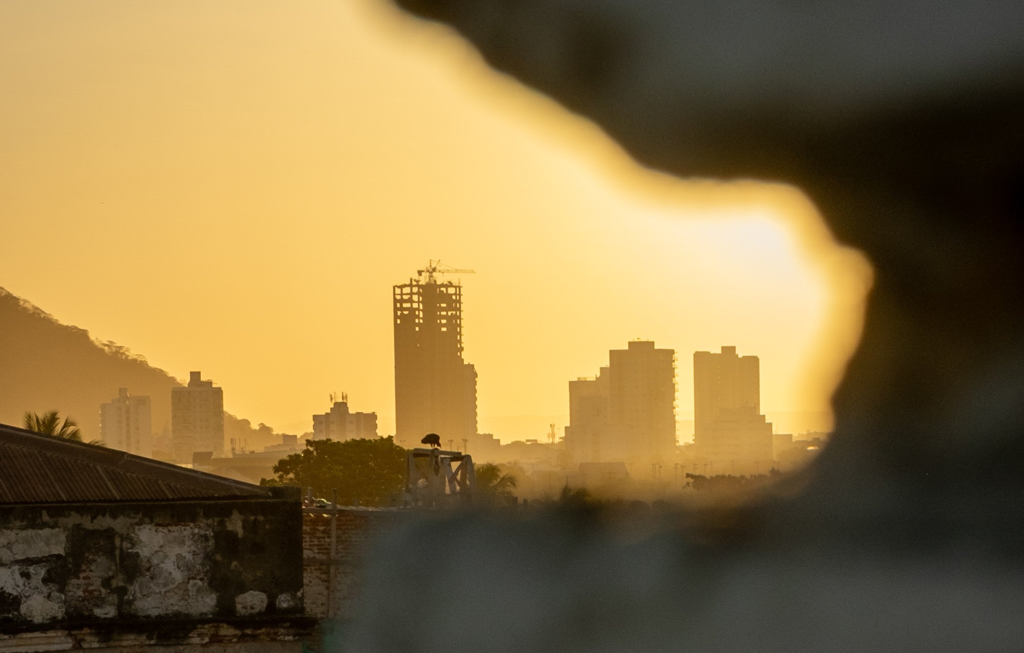 City skyline at sunset or sunrise, partially obscured by a dark object in the foreground, with several high-rise buildings and a crane visible.