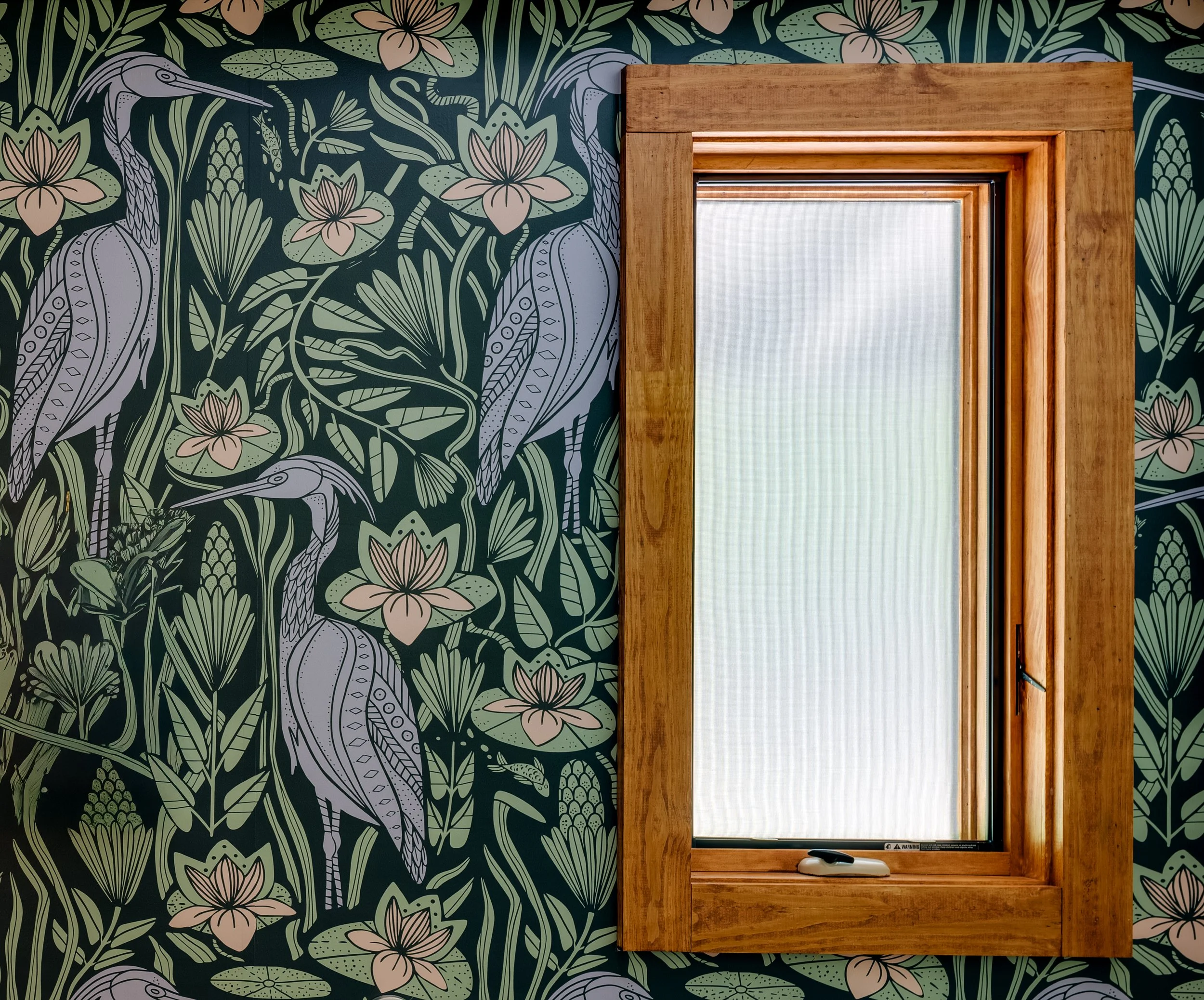 A window with a wooden frame in front of wallpaper with a botanical design featuring flowers, leaves, and a bird.