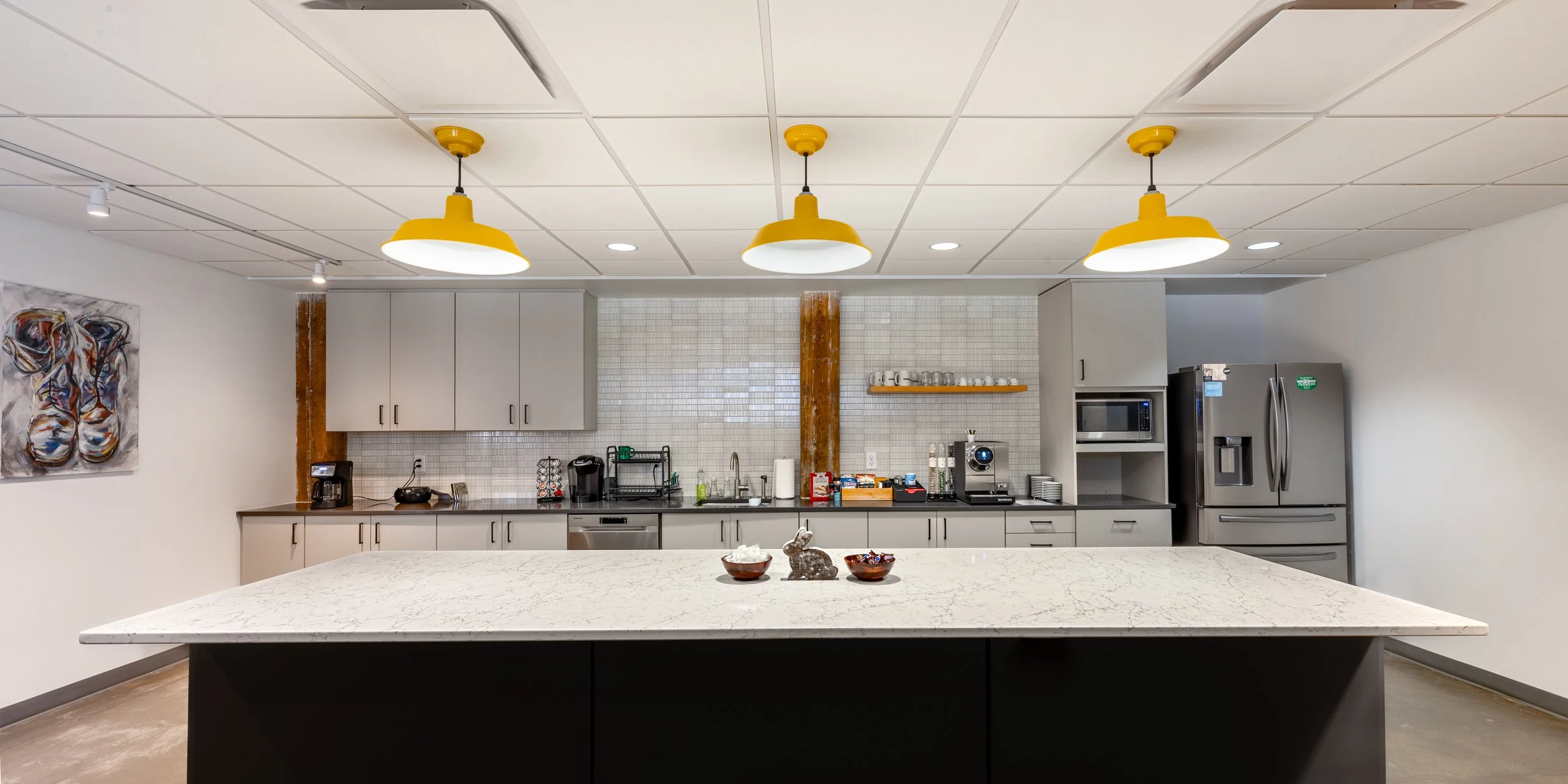 Modern kitchen with a large white marble island, gray cabinets, stainless steel appliances, yellow pendant lights, and abstract painting on the wall.