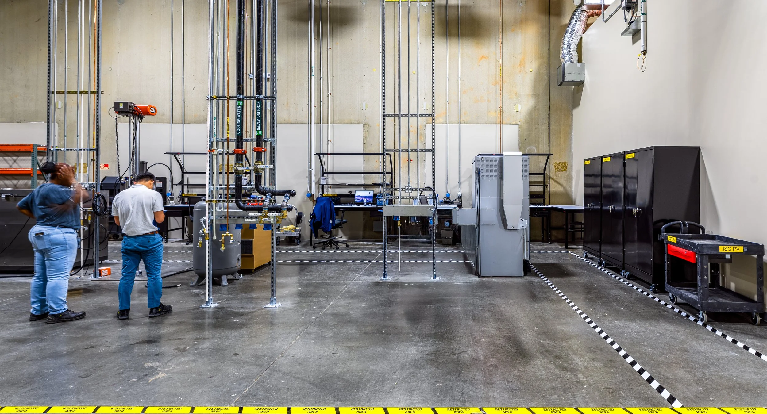 Two people working in a laboratory with various equipment, pipes, and tools, with a marked off area on the floor.