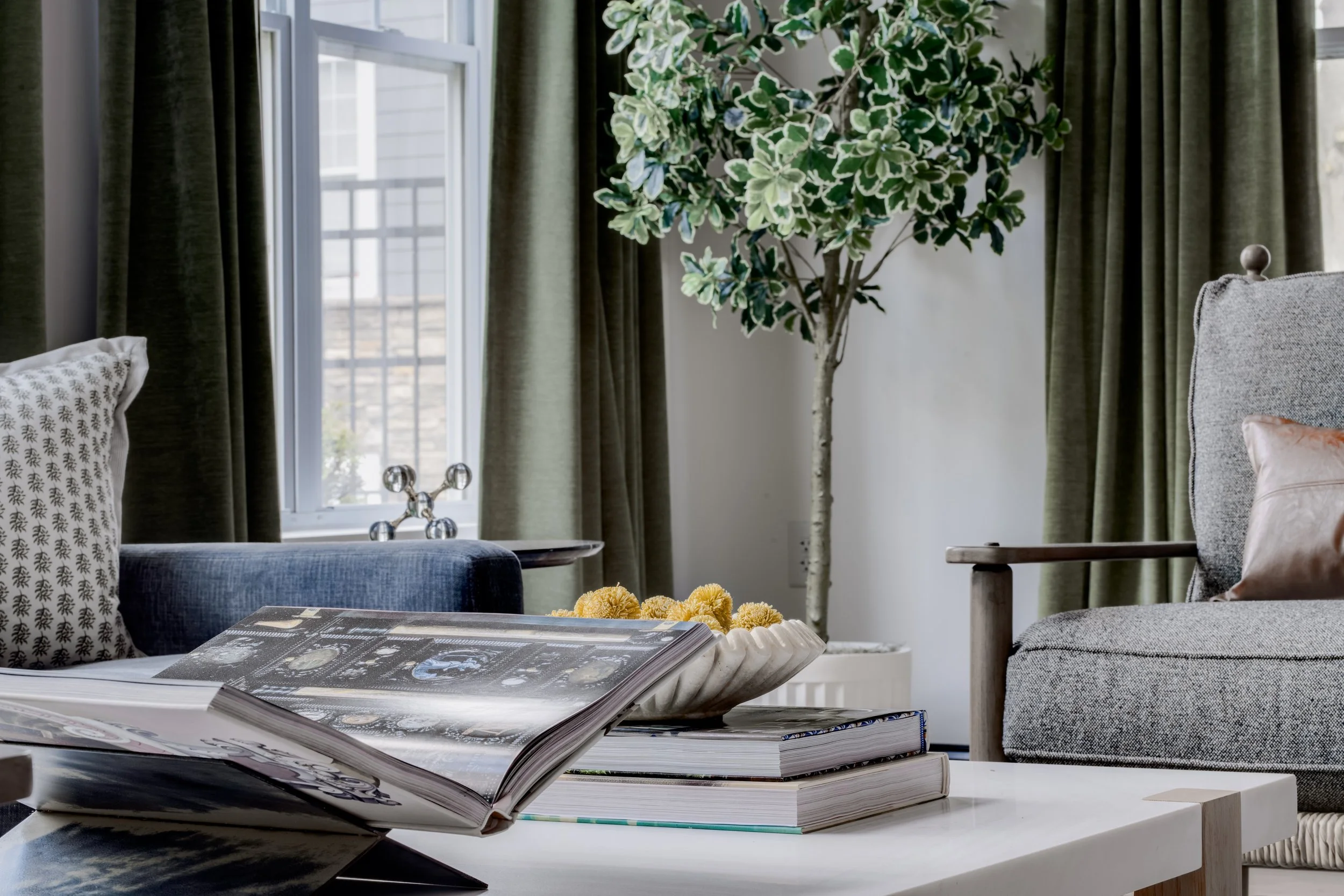A living room with a coffee table, open magazine, and decorative items, with a view of window and green curtains, and a large potted plant in the background.