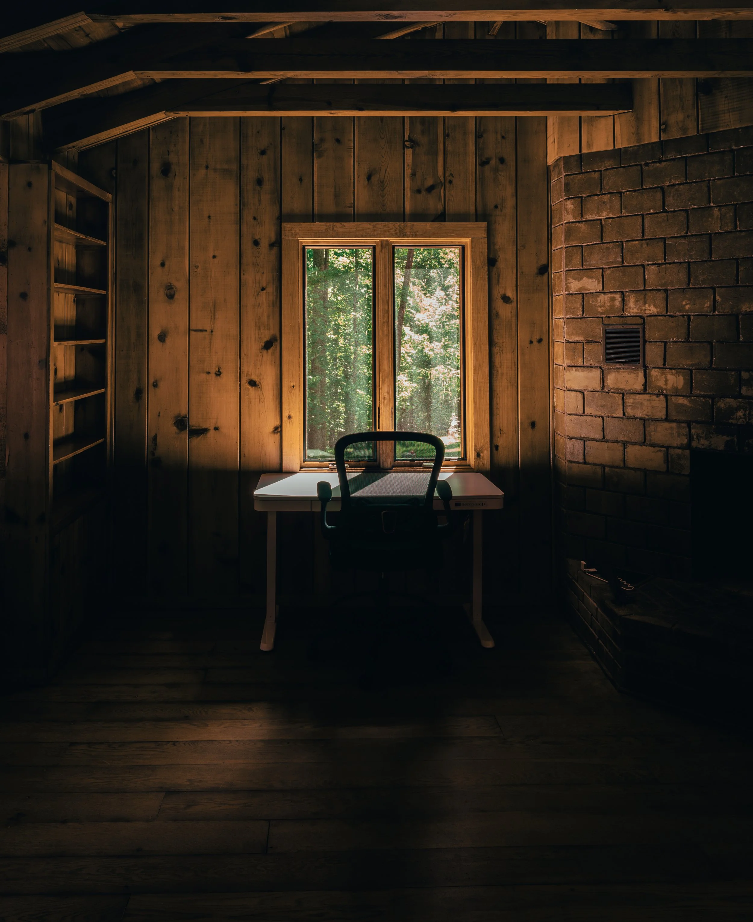 A wooden interior room with vertical wood panel walls, a brick chimney on the right, and a window showing green trees outside. A desk and chair are in front of the window.