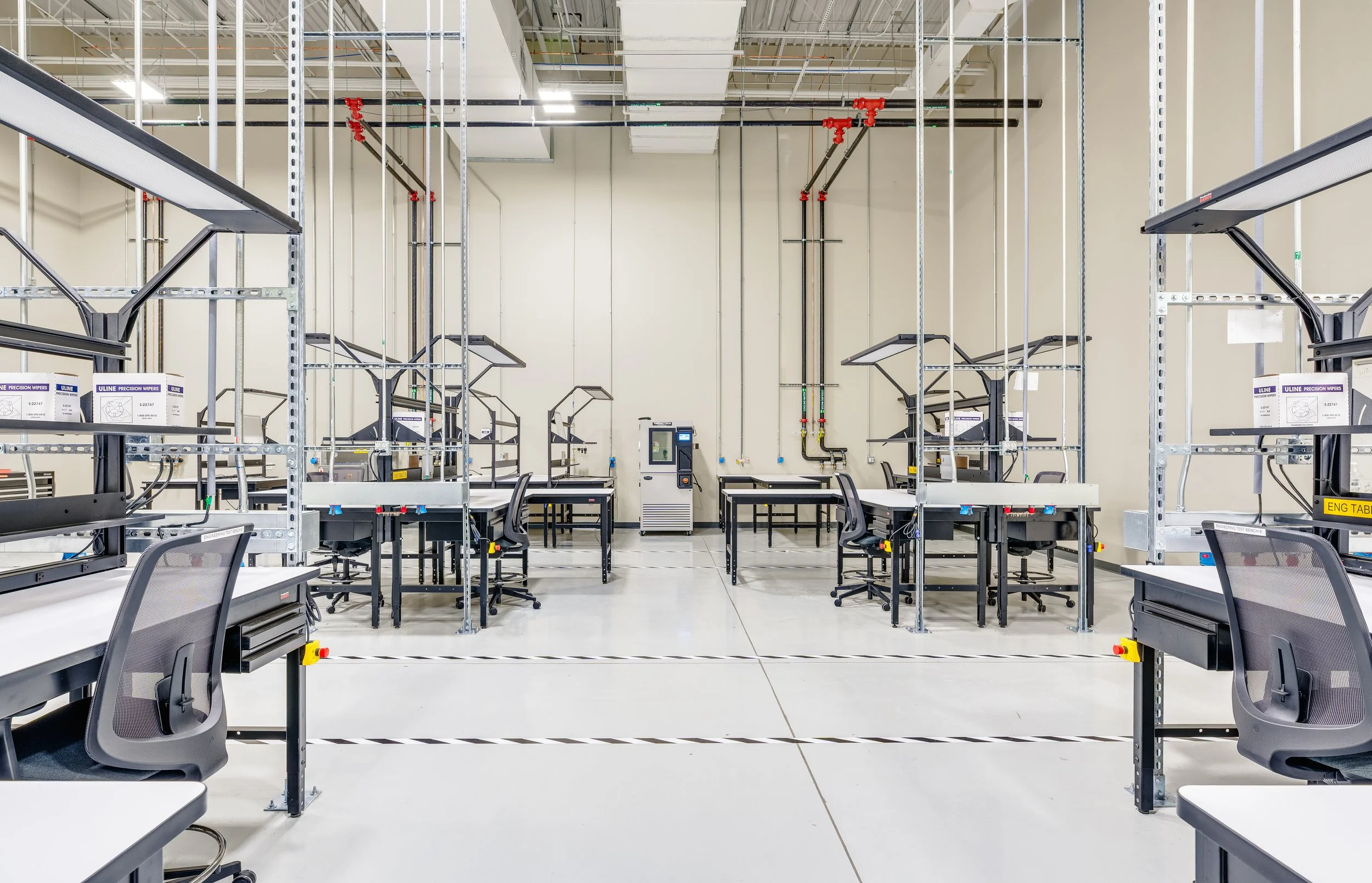 Empty industrial workspace with metal shelving, work tables, chairs, and electrical conduits on the walls.